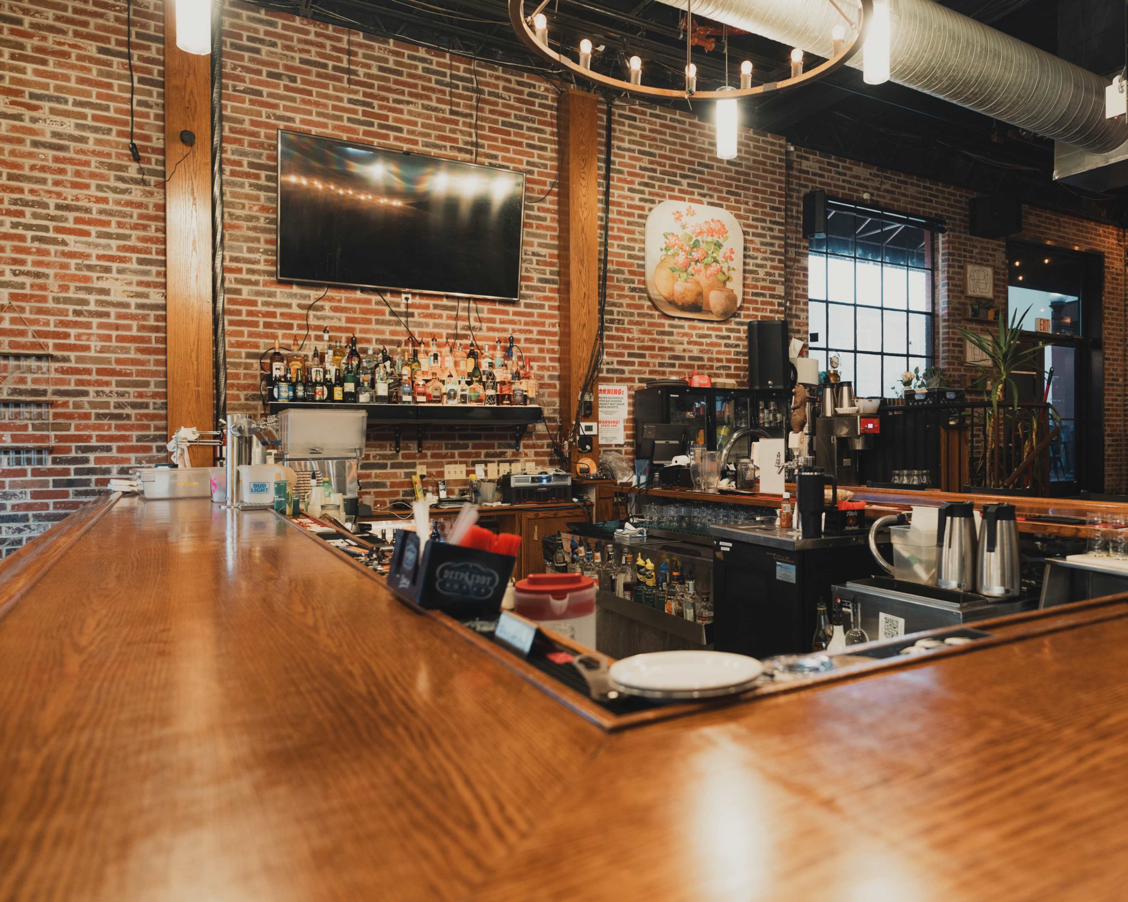 The image shows the interior of a bar with a wooden countertop, a variety of liquor bottles on shelves, and exposed brick walls decorated with artwork.