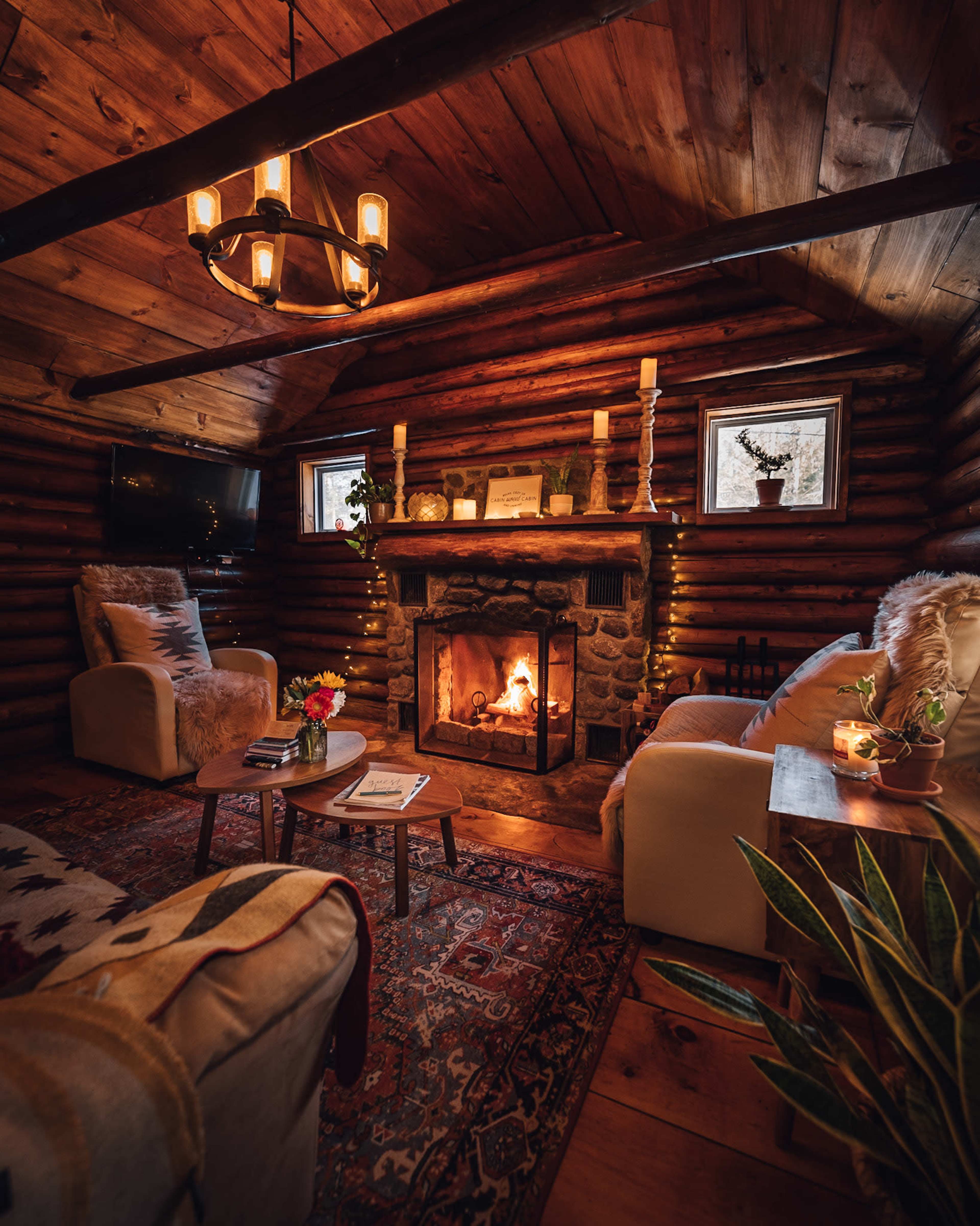 The interior of a cozy log cabin features a stone fireplace, two armchairs, a small table with magazines, and decorative plants.
