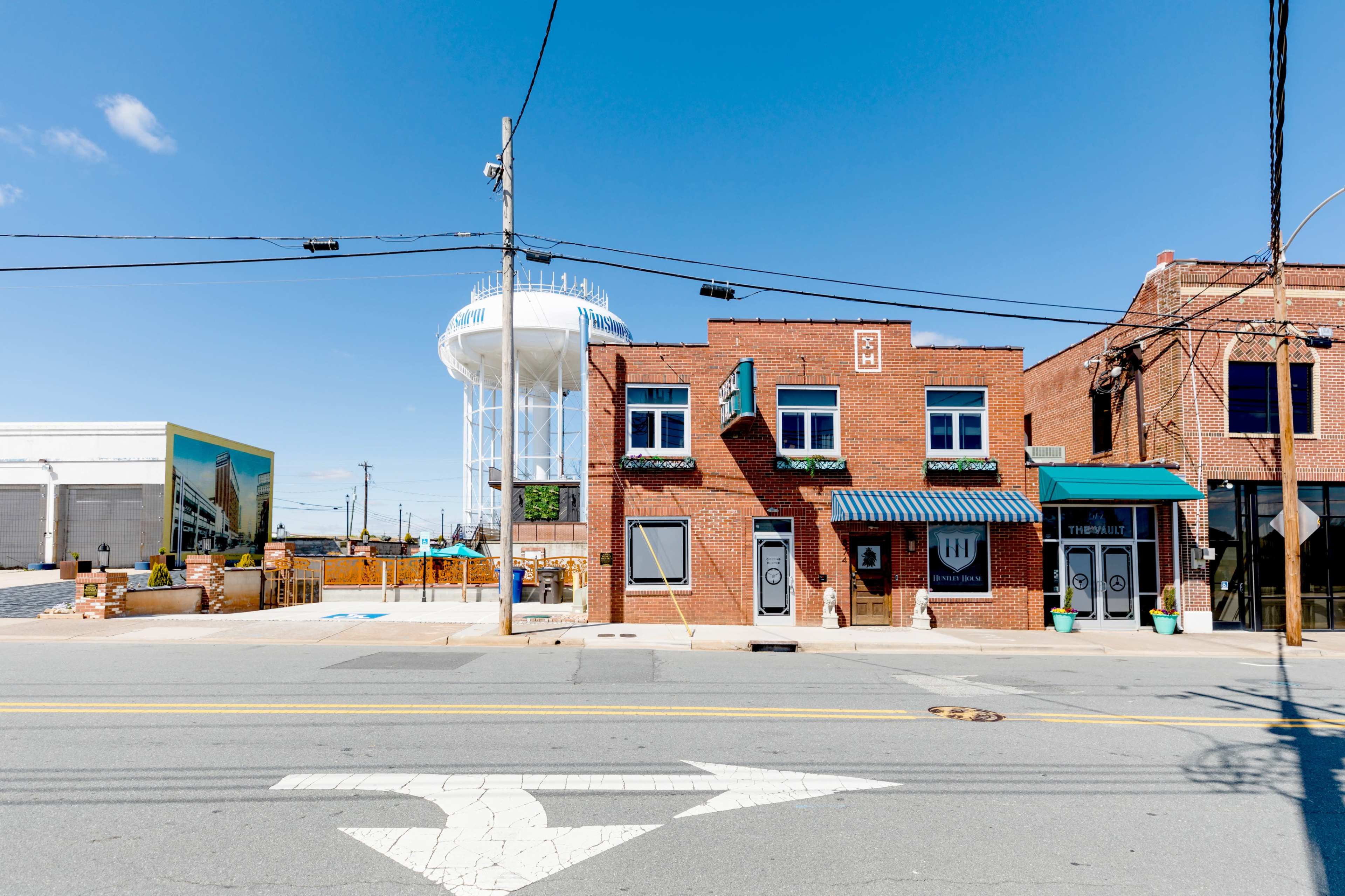 A row of brick buildings with awnings, a street sign directing traffic, and a water tower in the background against a clear blue sky.