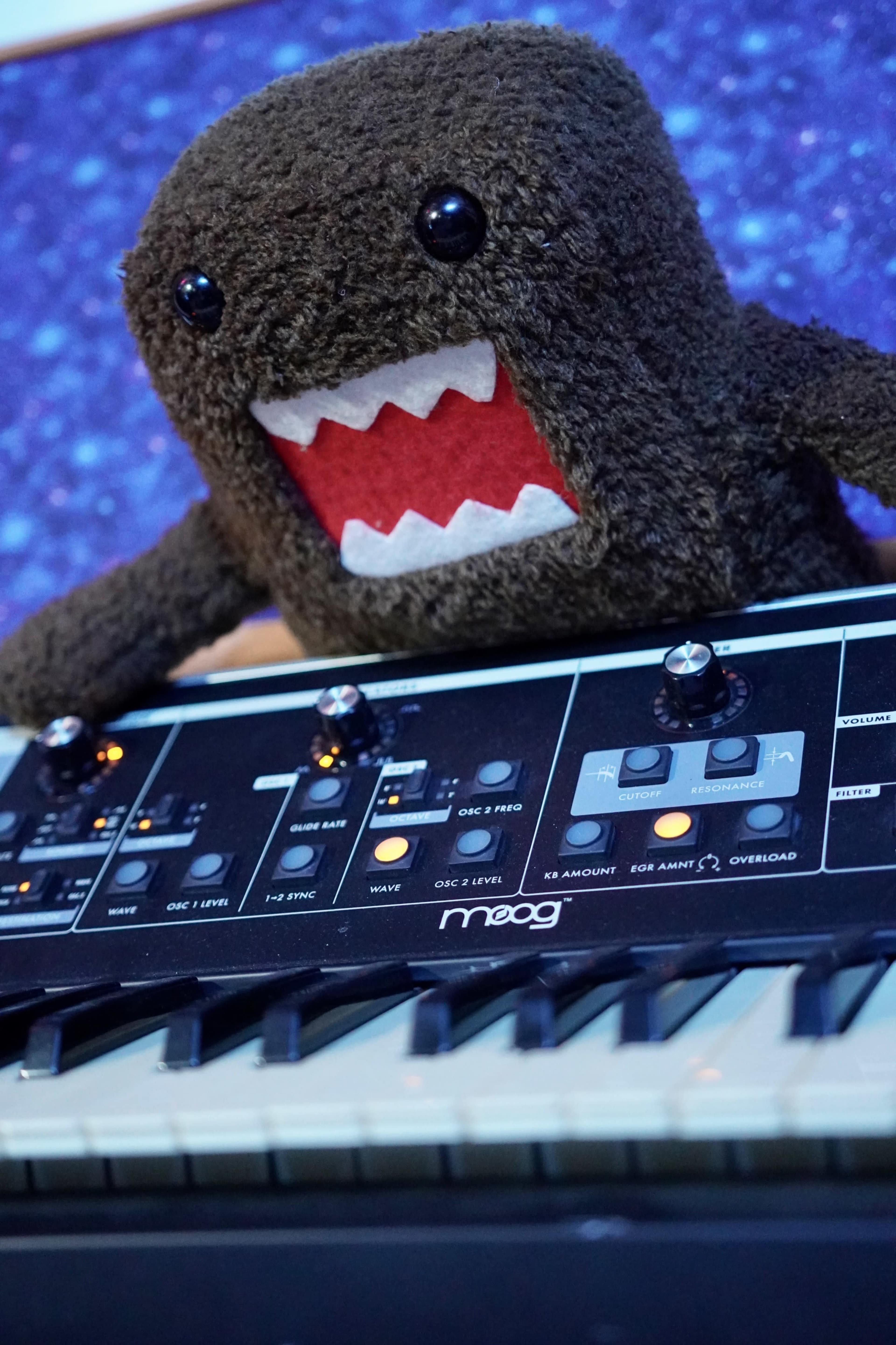 A plush toy with a wide mouth sits atop a synthesizer keyboard, featuring various knobs and buttons.