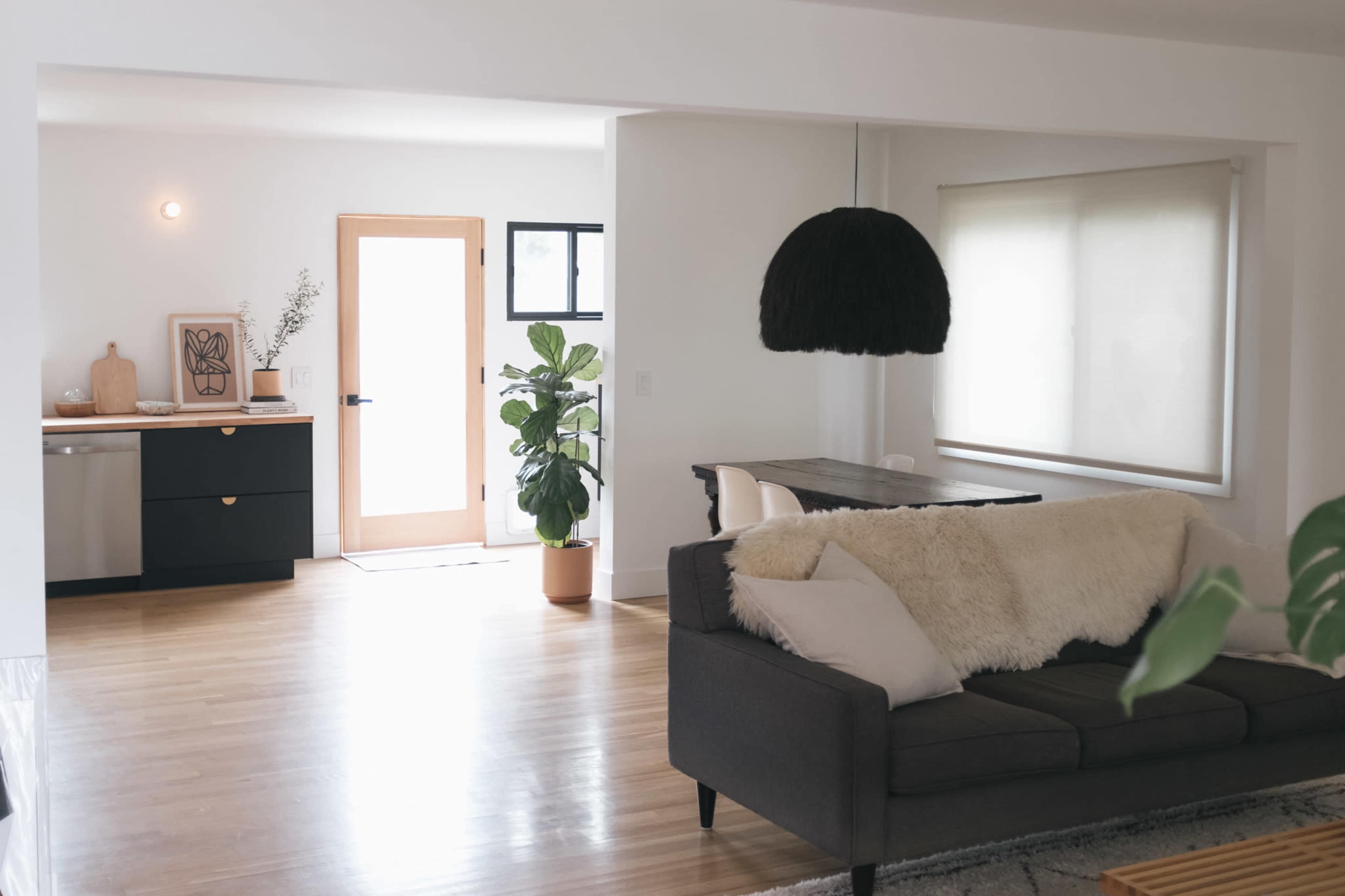 A modern living space features a grey couch with a white throw, a dining area with a wooden table, and a door leading to the outside, illuminated by natural light.
