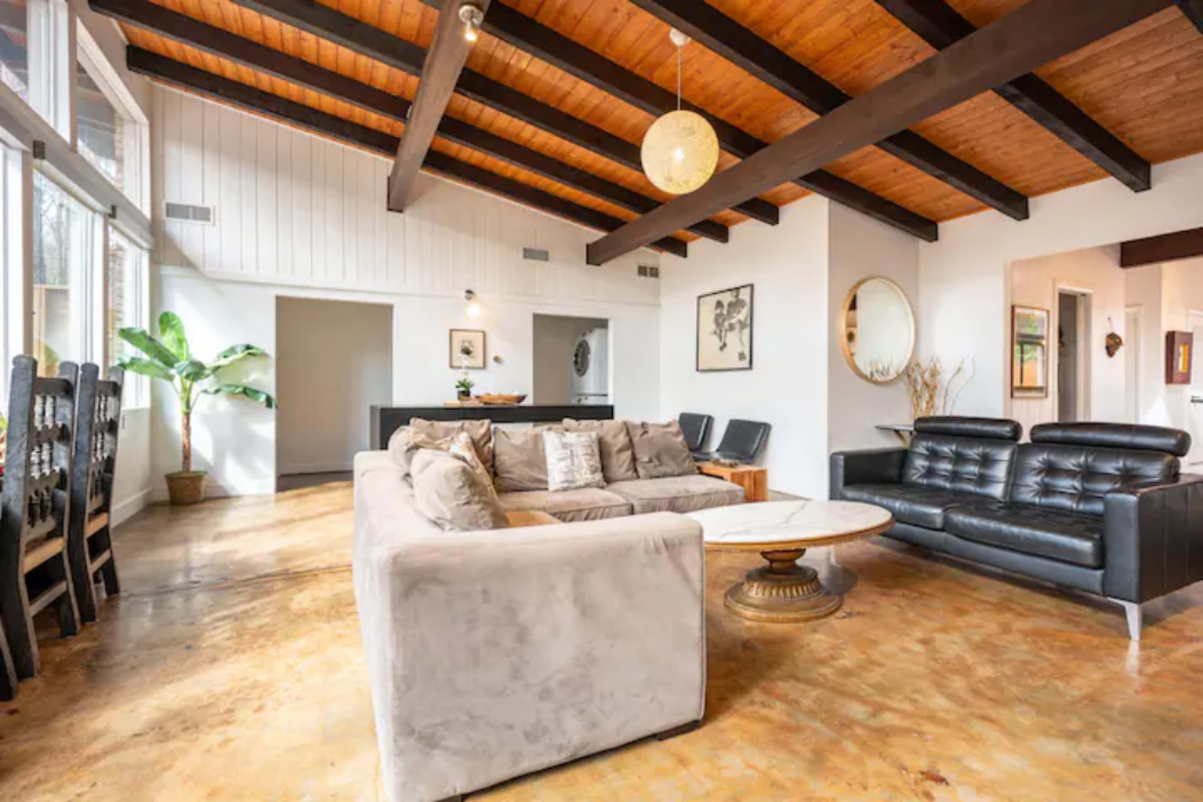The image shows a modern living room with a high ceiling, exposed wooden beams, a beige sectional sofa, black leather chairs, and a round coffee table.