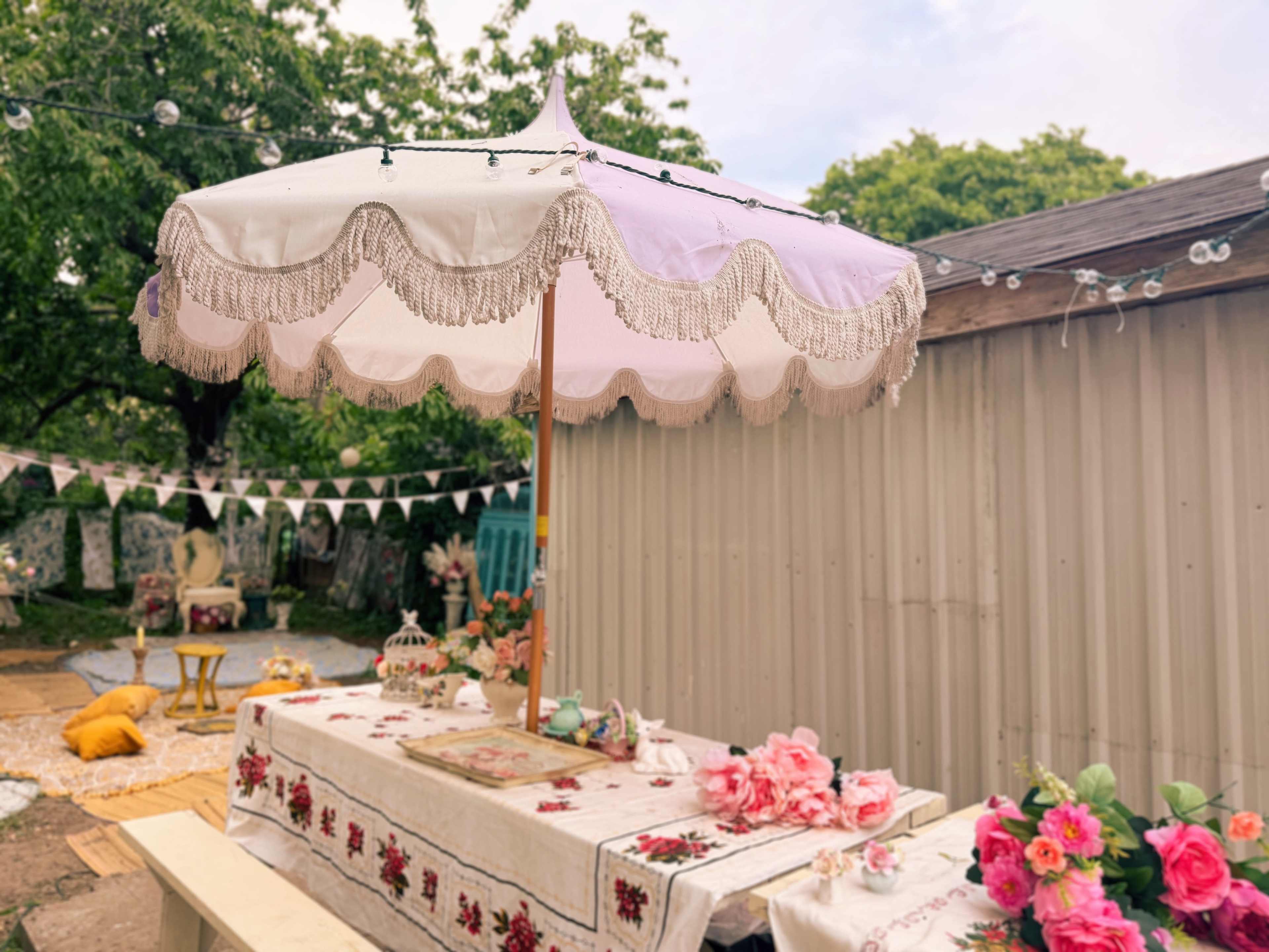 A decorated outdoor space features a large umbrella with decorative fringe over a table adorned with flowers and various decorative items.