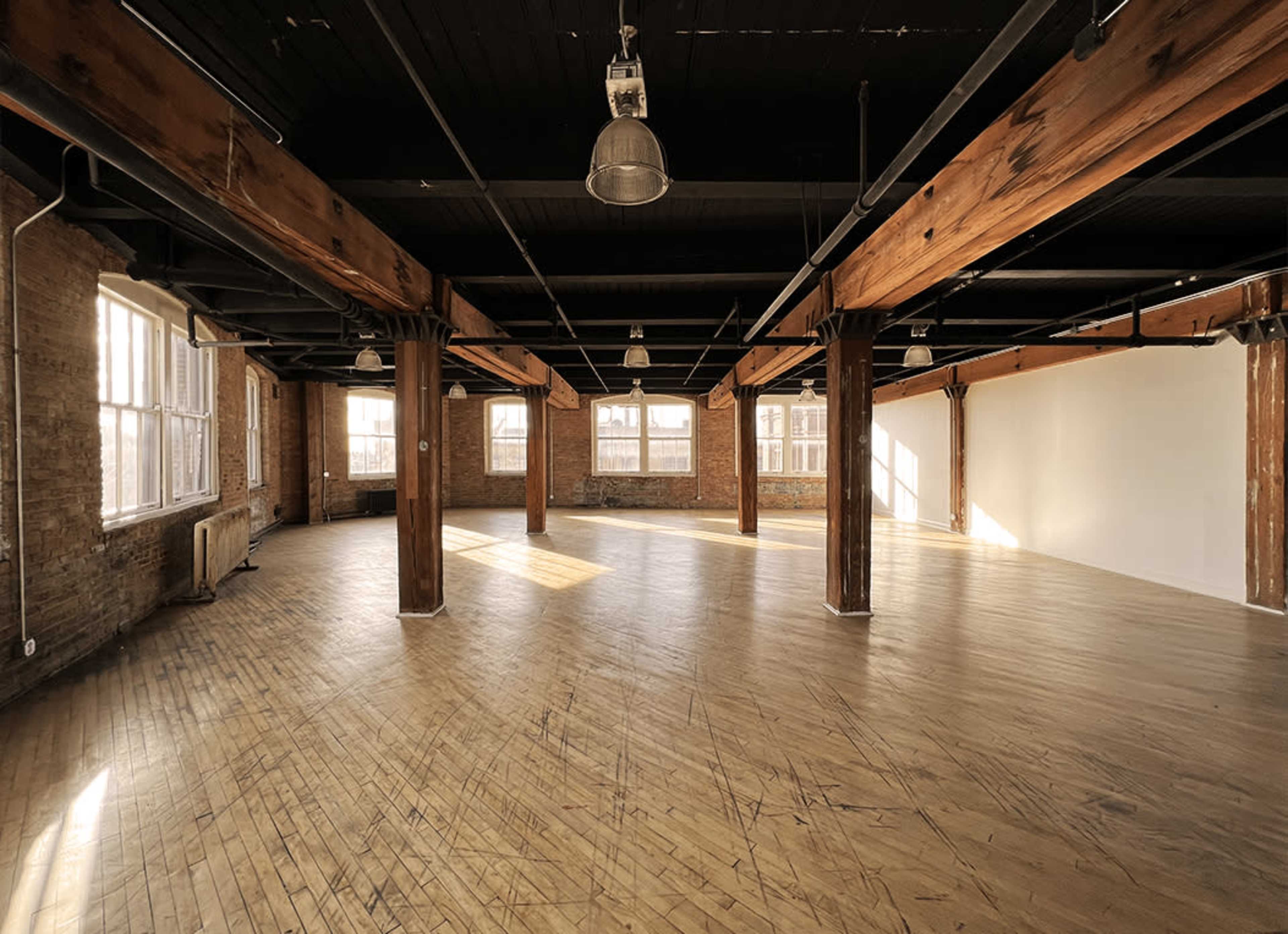 A spacious, empty room with wooden floors and large windows, accented by exposed beams and pillars.