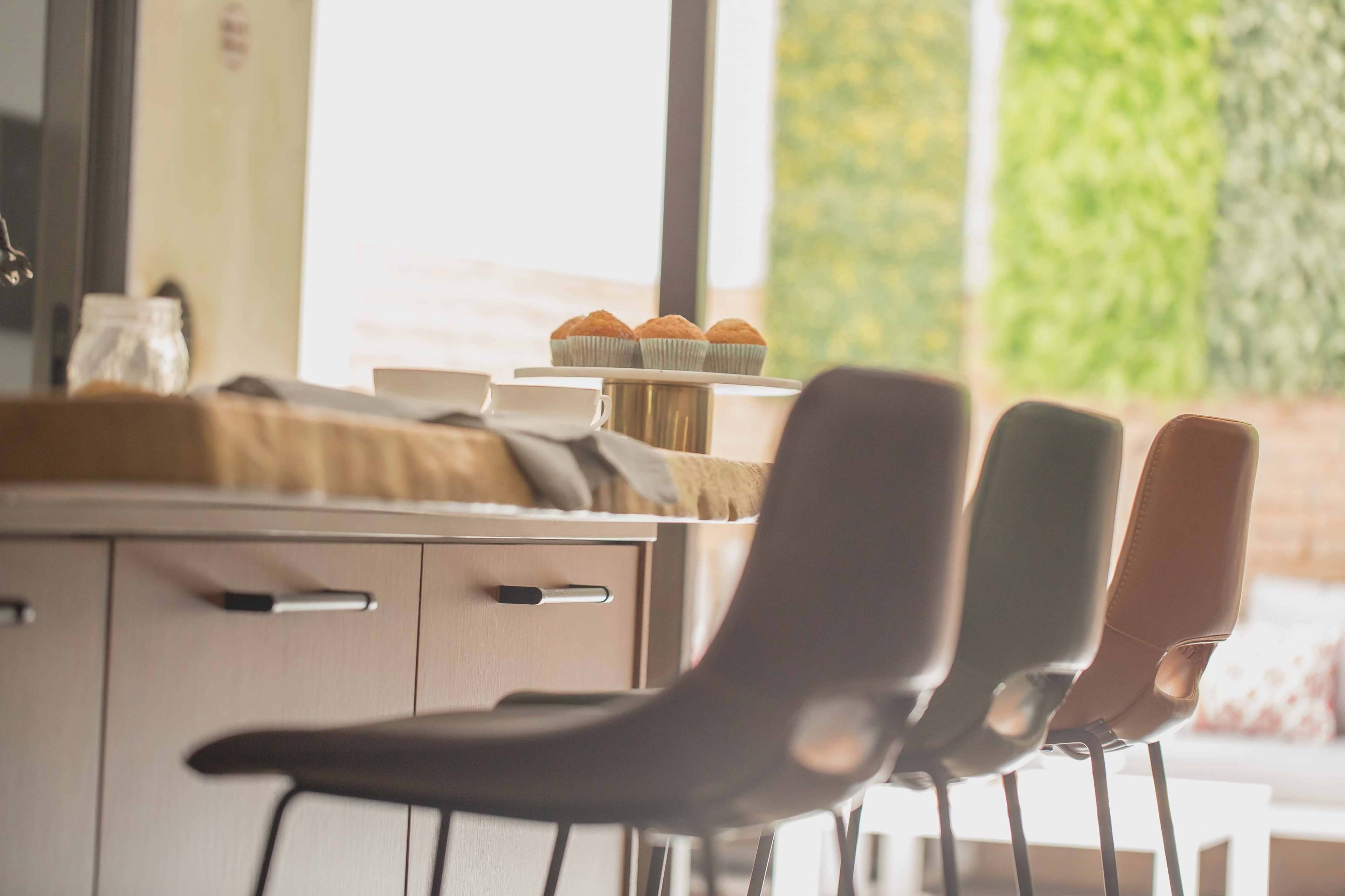 A kitchen counter is set with a plate of cupcakes, three bar stools lined up nearby, and greenery visible through the window.