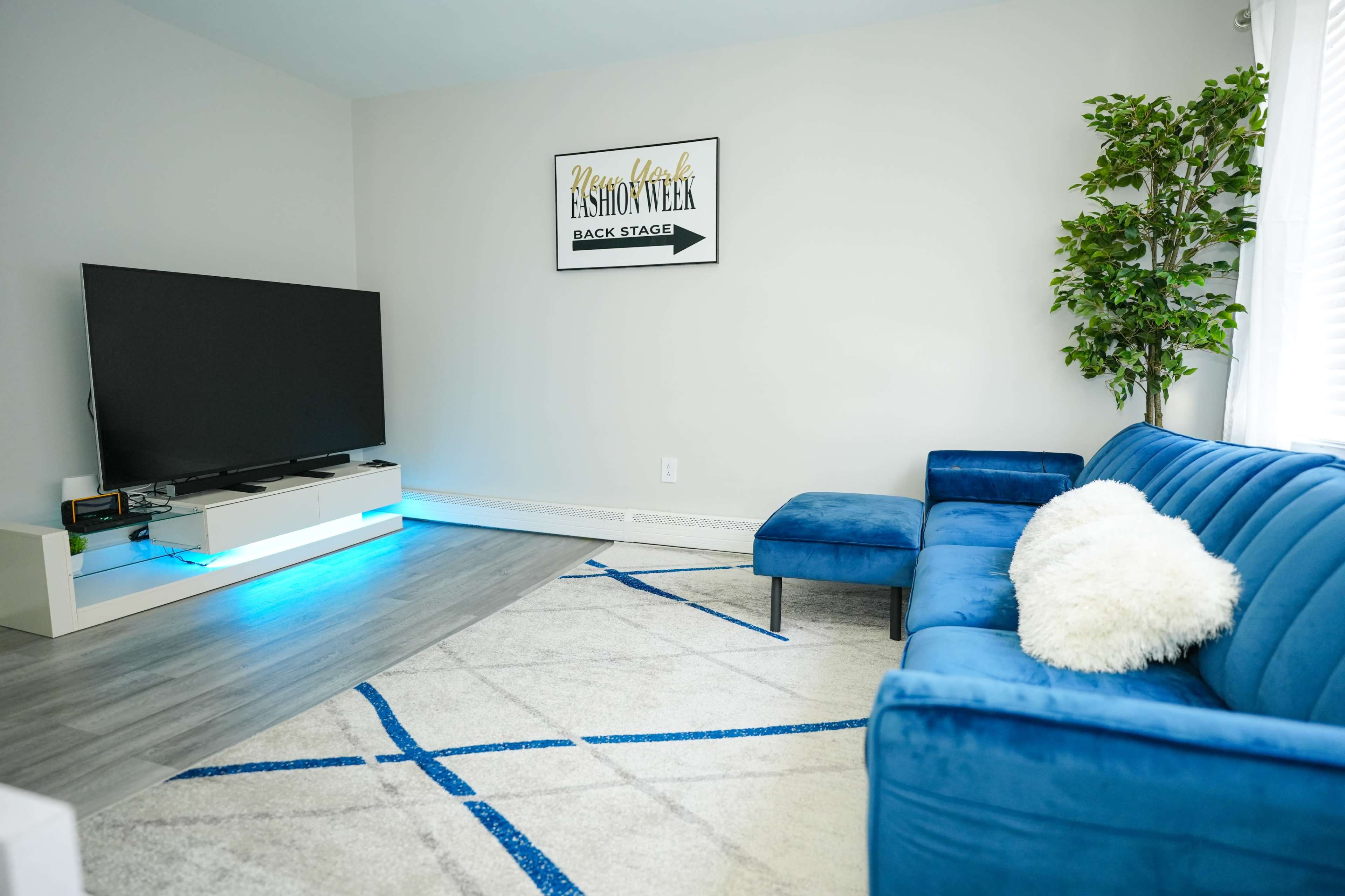 A modern living room with a blue sectional sofa, a large television on a white stand, and a decorative plant beside a light-colored rug.
