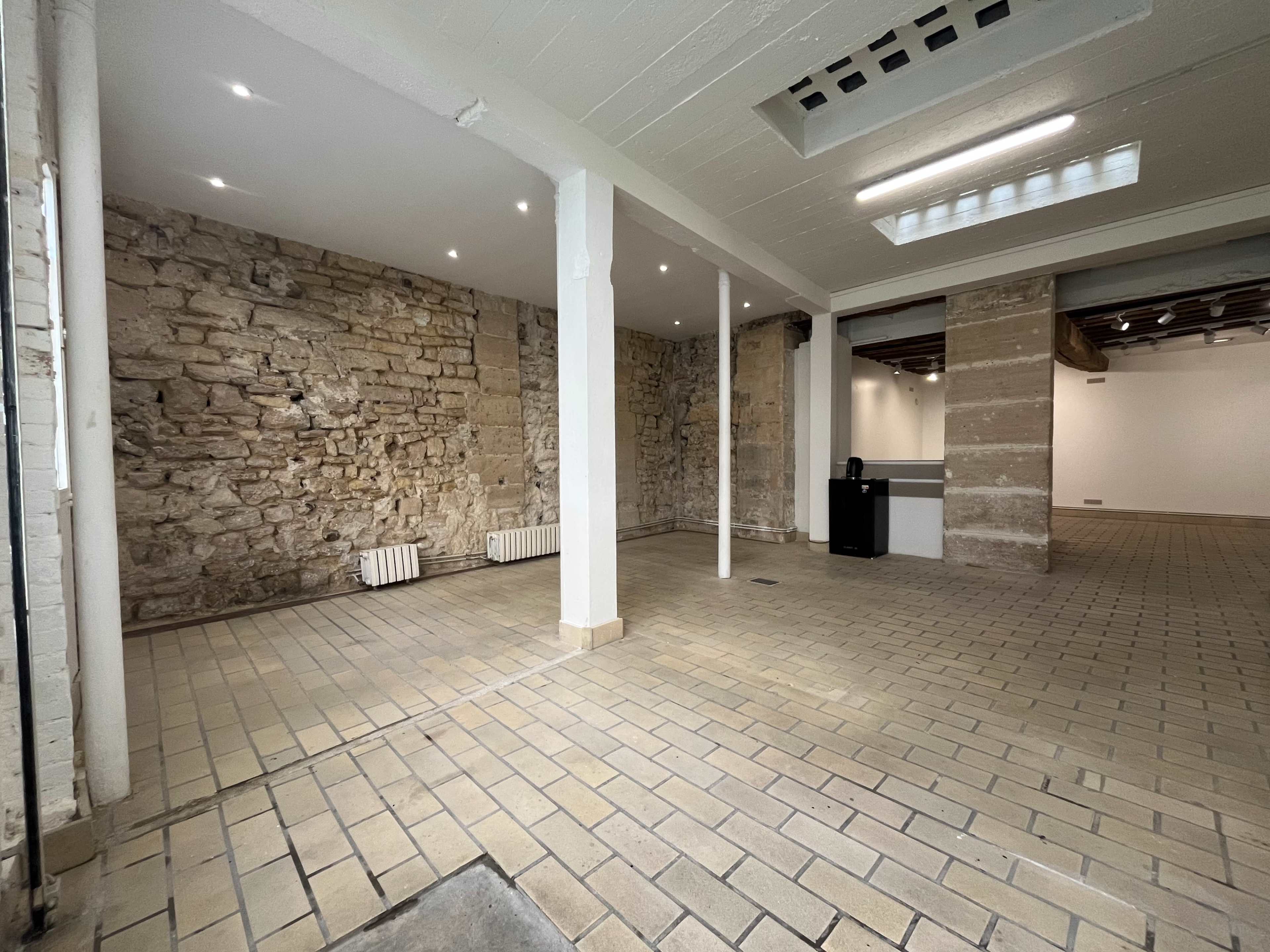 The image shows a spacious, well-lit interior with stone walls and a tiled floor, featuring a few columns and a small kitchenette area in one corner.