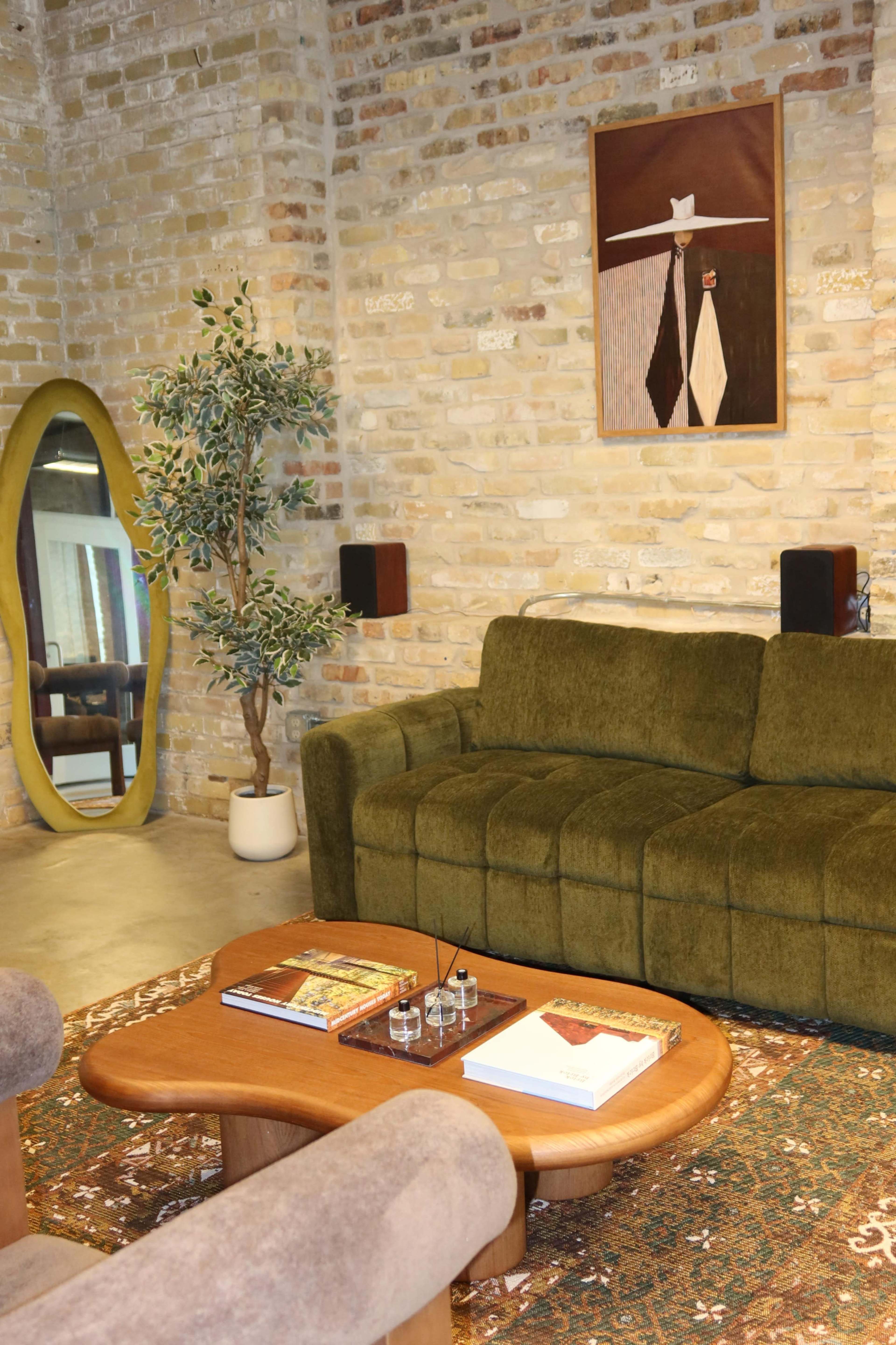 The image shows a cozy living room with a green sofa, a wooden coffee table, a potted plant, and a large round mirror against a brick wall.