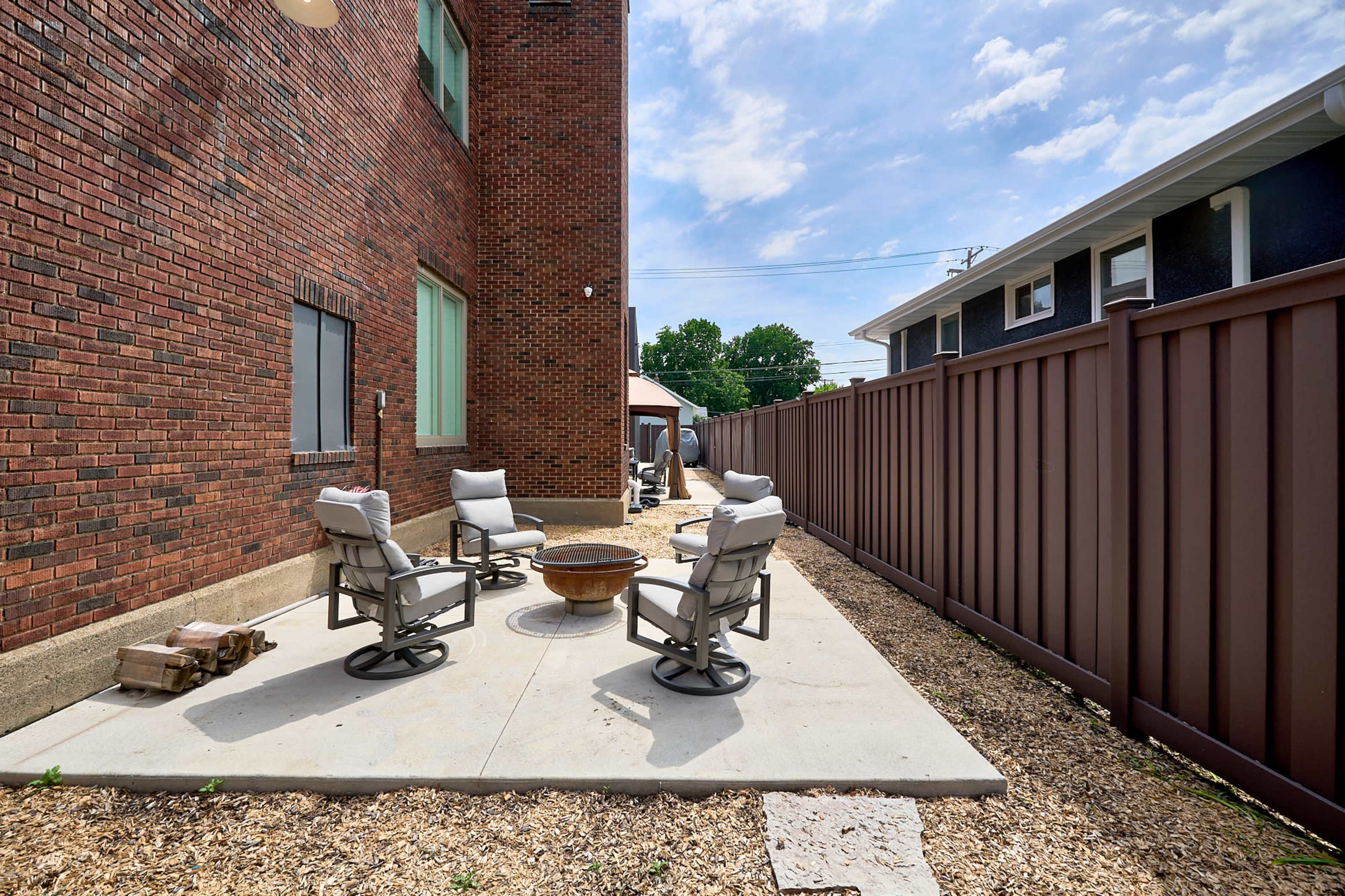A small outdoor seating area features four swivel chairs arranged around a fire pit, enclosed by a wooden fence and adjacent to a brick building.