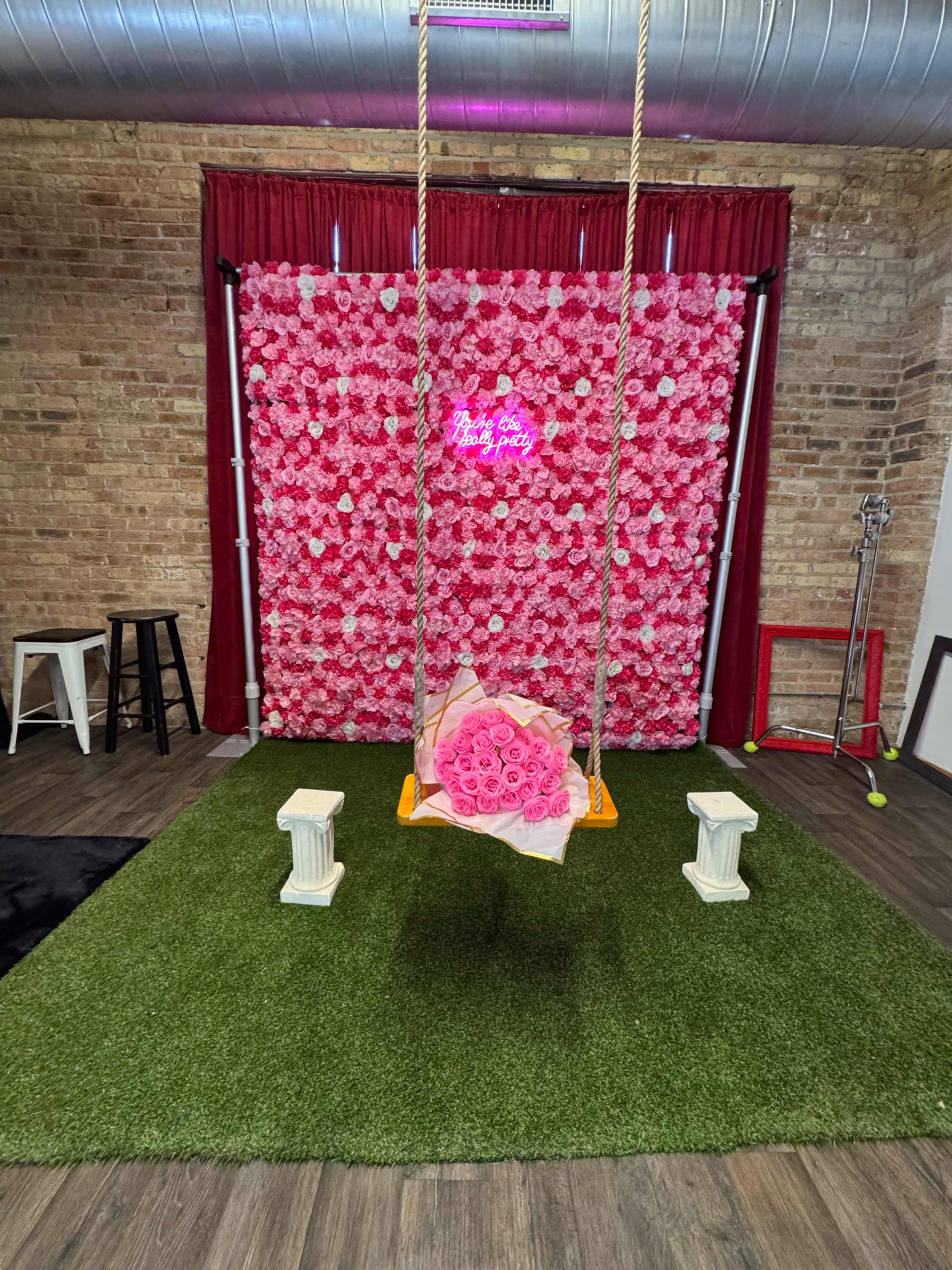 The image shows a decorative setup featuring a swing in front of a pink floral wall, with a bouquet of roses on the ground and green grass-like carpet underneath.