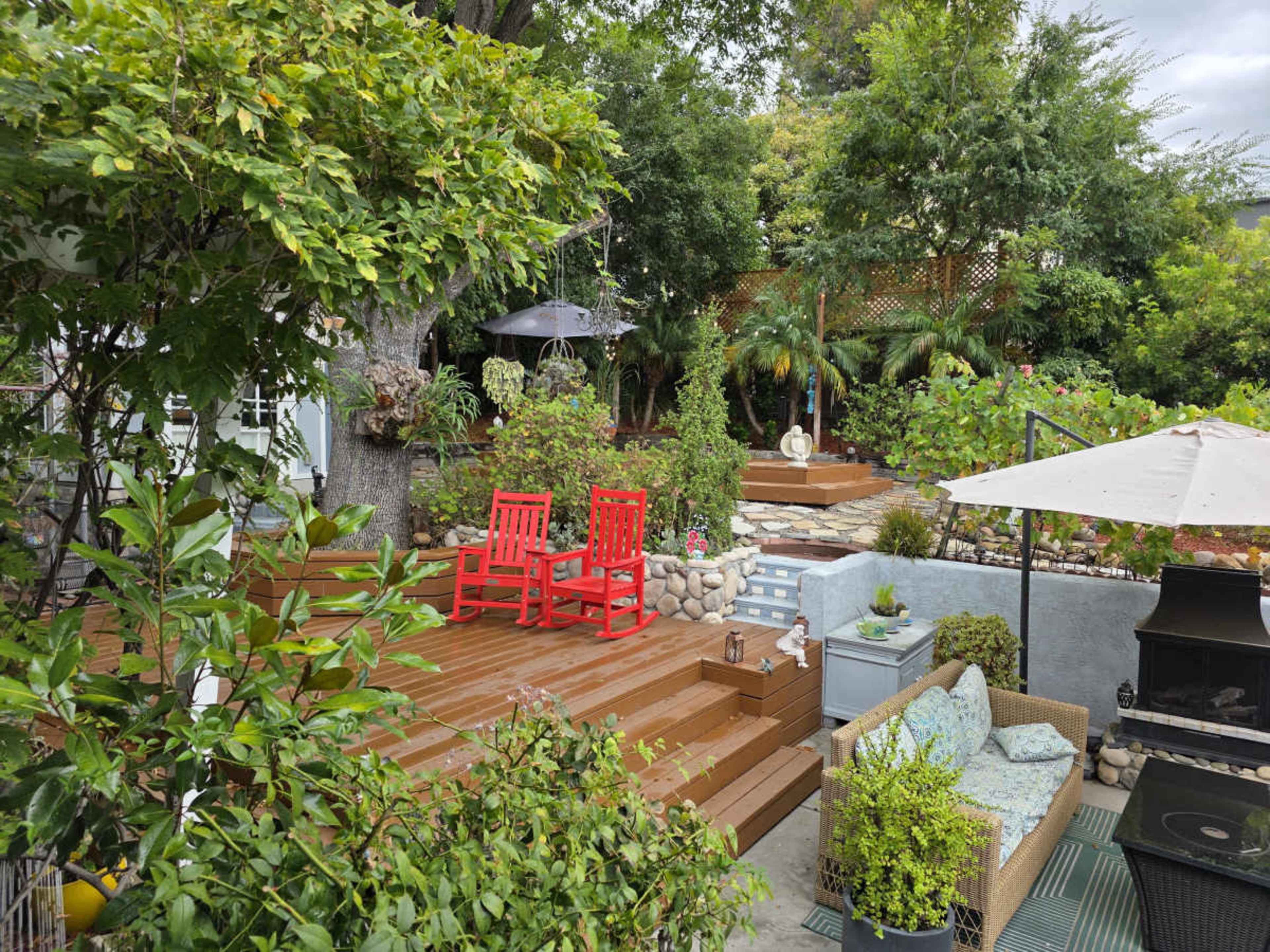 The image shows a landscaped backyard featuring a wooden deck with two red chairs, surrounded by various plants and outdoor furniture.