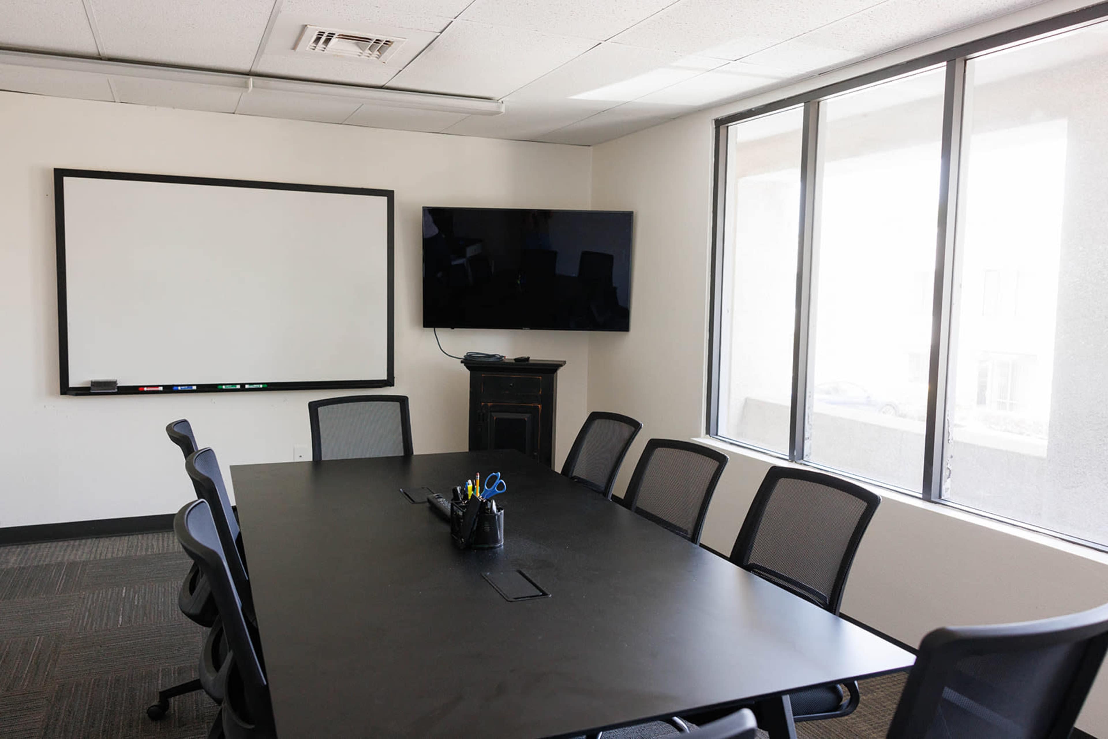 The image shows a modern conference room featuring a large black table, ergonomic chairs, a whiteboard, and a mounted television, all illuminated by natural light from the windows.
