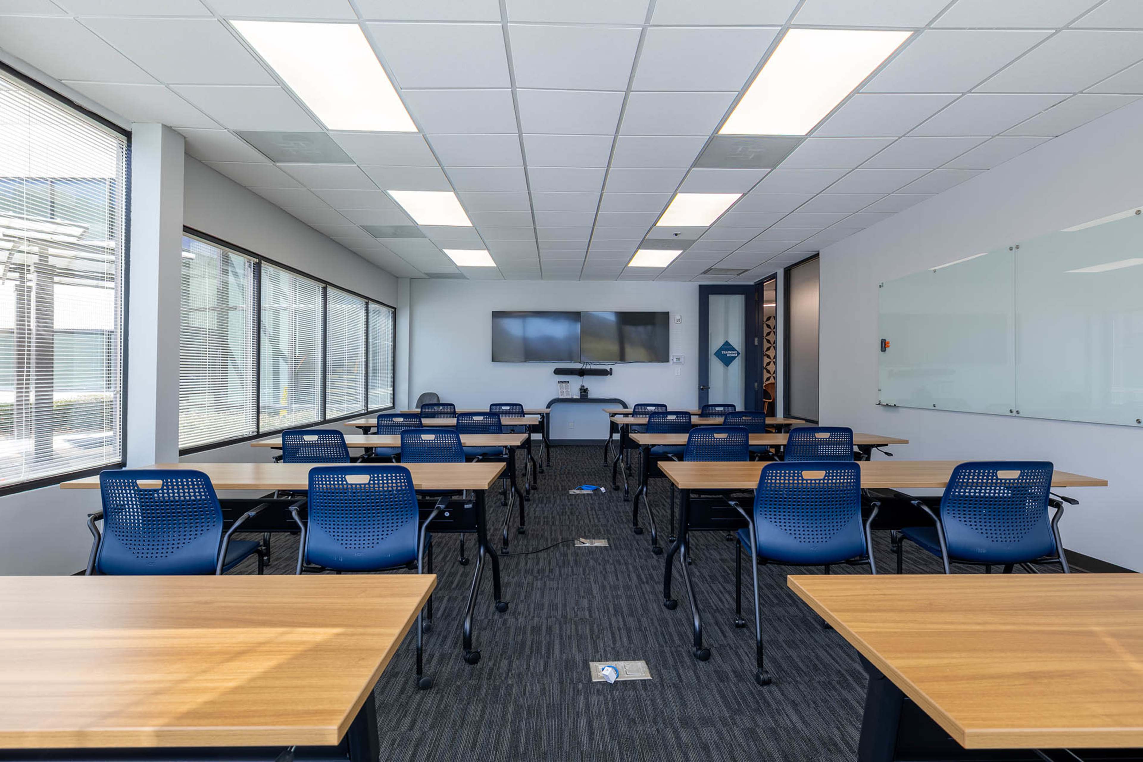 The image shows a modern classroom with several rows of wooden tables and blue chairs, equipped with a wall-mounted screen and whiteboards.