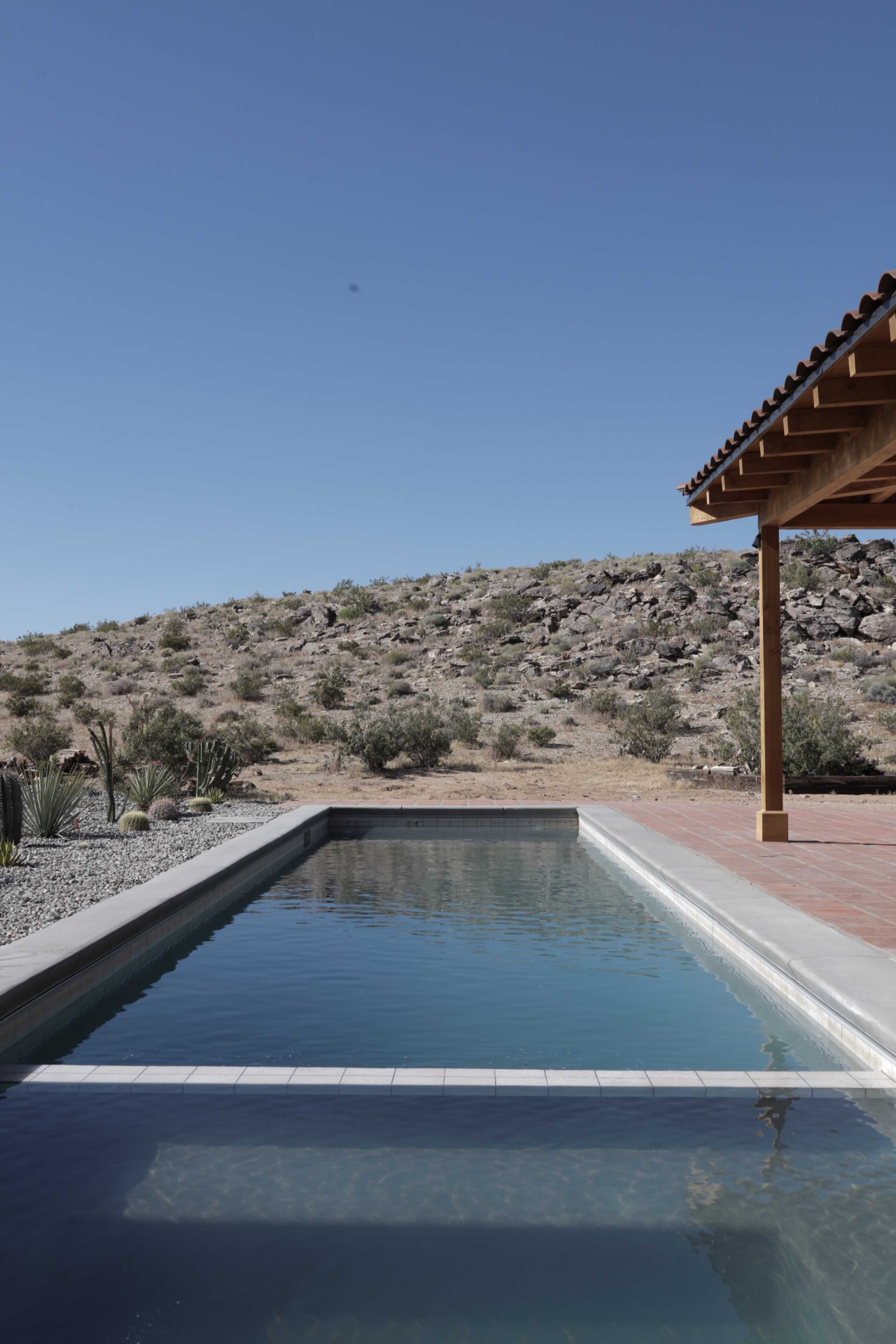 A rectangular swimming pool is positioned next to a rocky hillside under a clear blue sky.