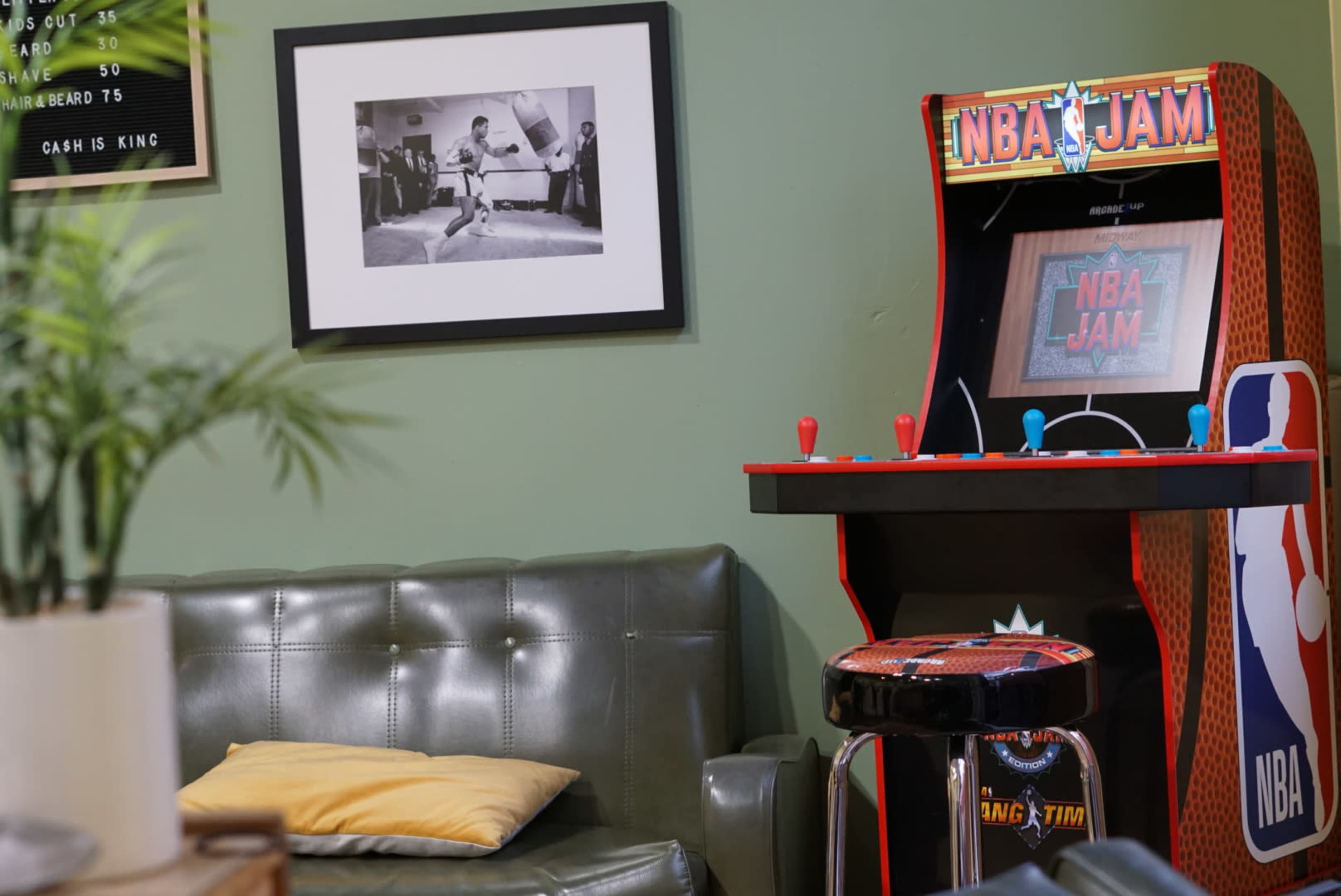 A retro NBA Jam arcade game next to a green leather sofa and a framed black-and-white photograph on the wall.