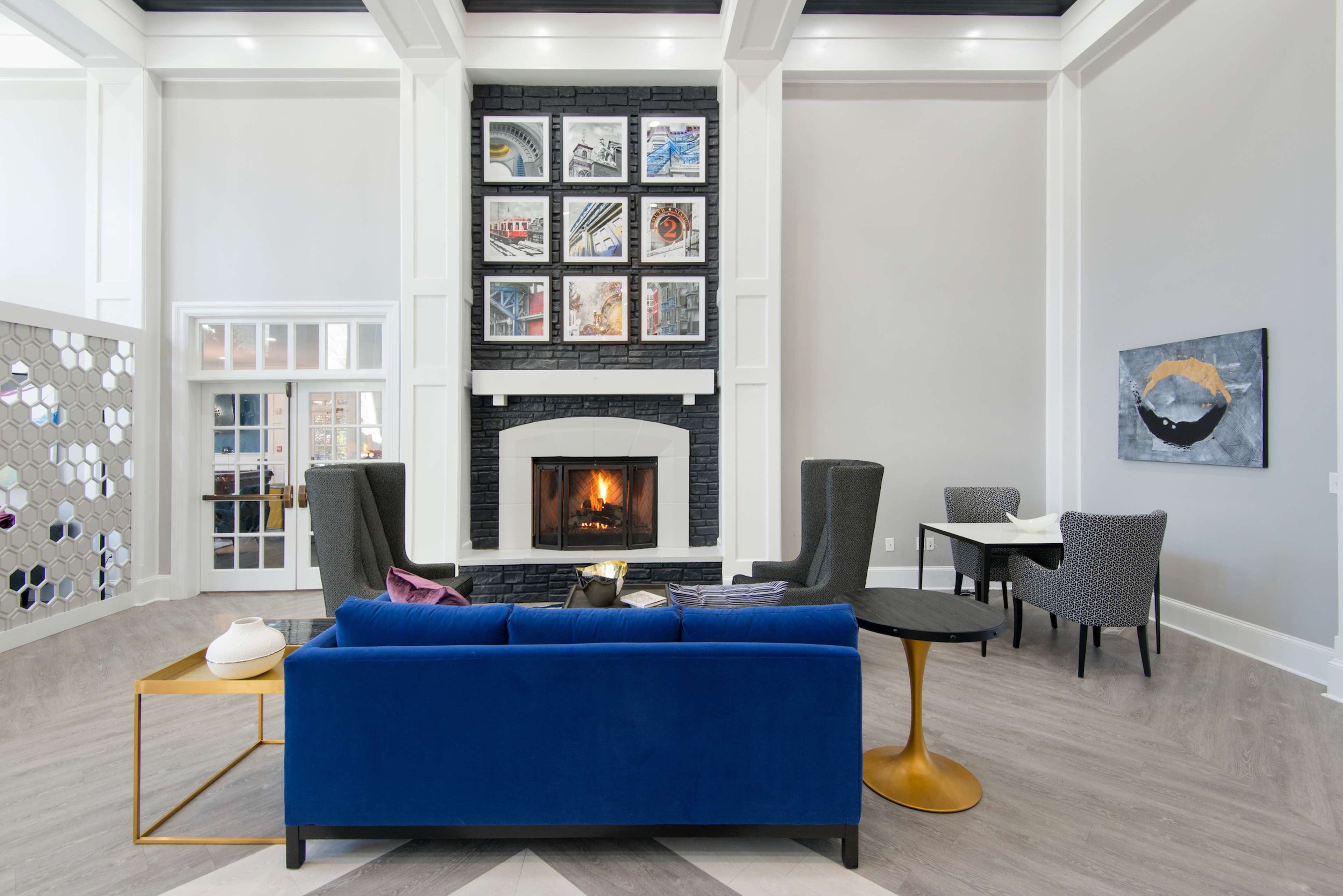 The image shows a modern lobby with a blue sofa, two high-backed chairs, a fireplace framed by a black stone wall, and a series of framed artworks above it.
