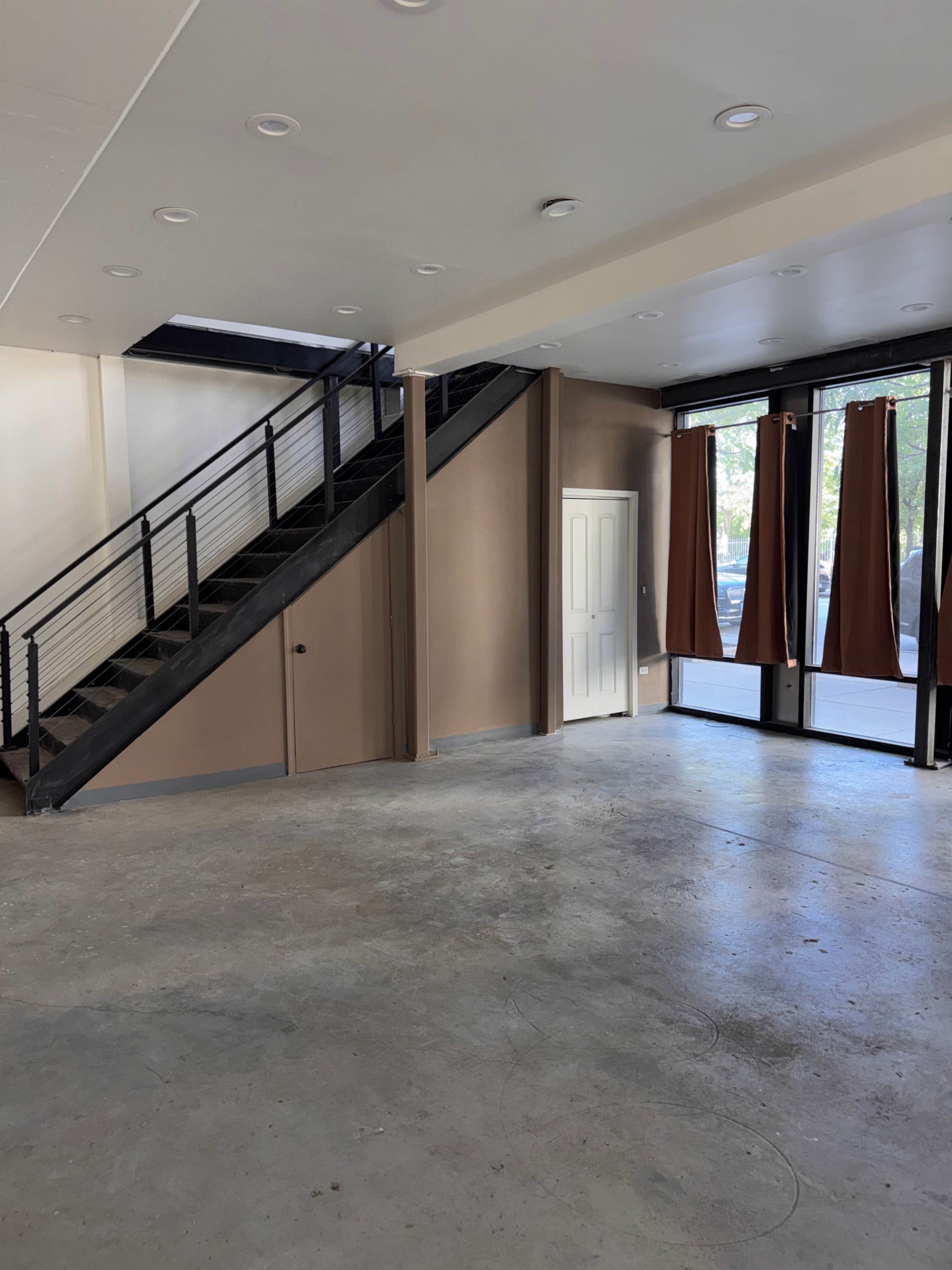 The image shows an empty room with concrete flooring, a staircase on the left, and large windows covered with brown curtains.