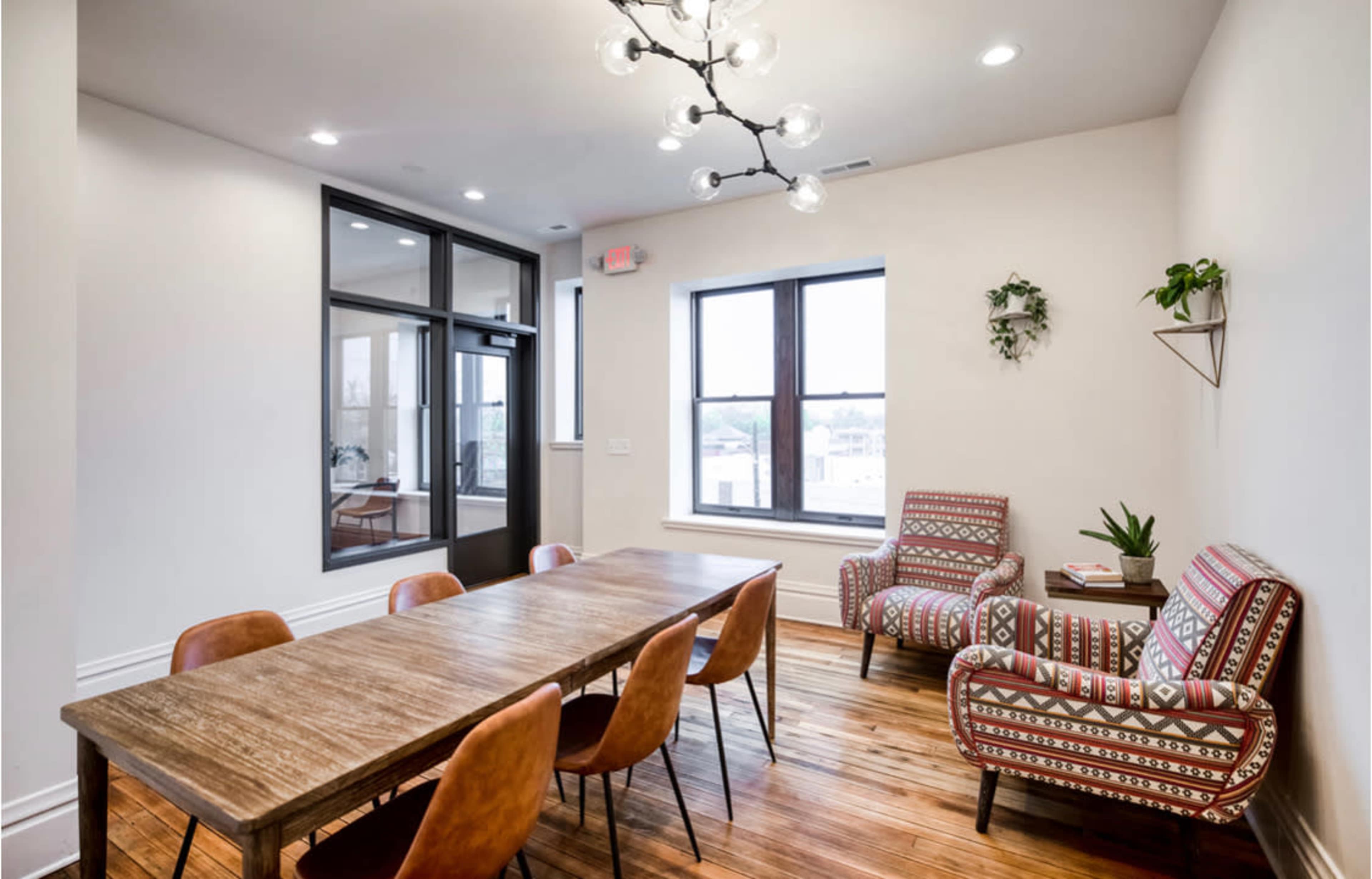 A meeting room features a long wooden table surrounded by brown chairs, with two patterned armchairs and large windows letting in natural light.