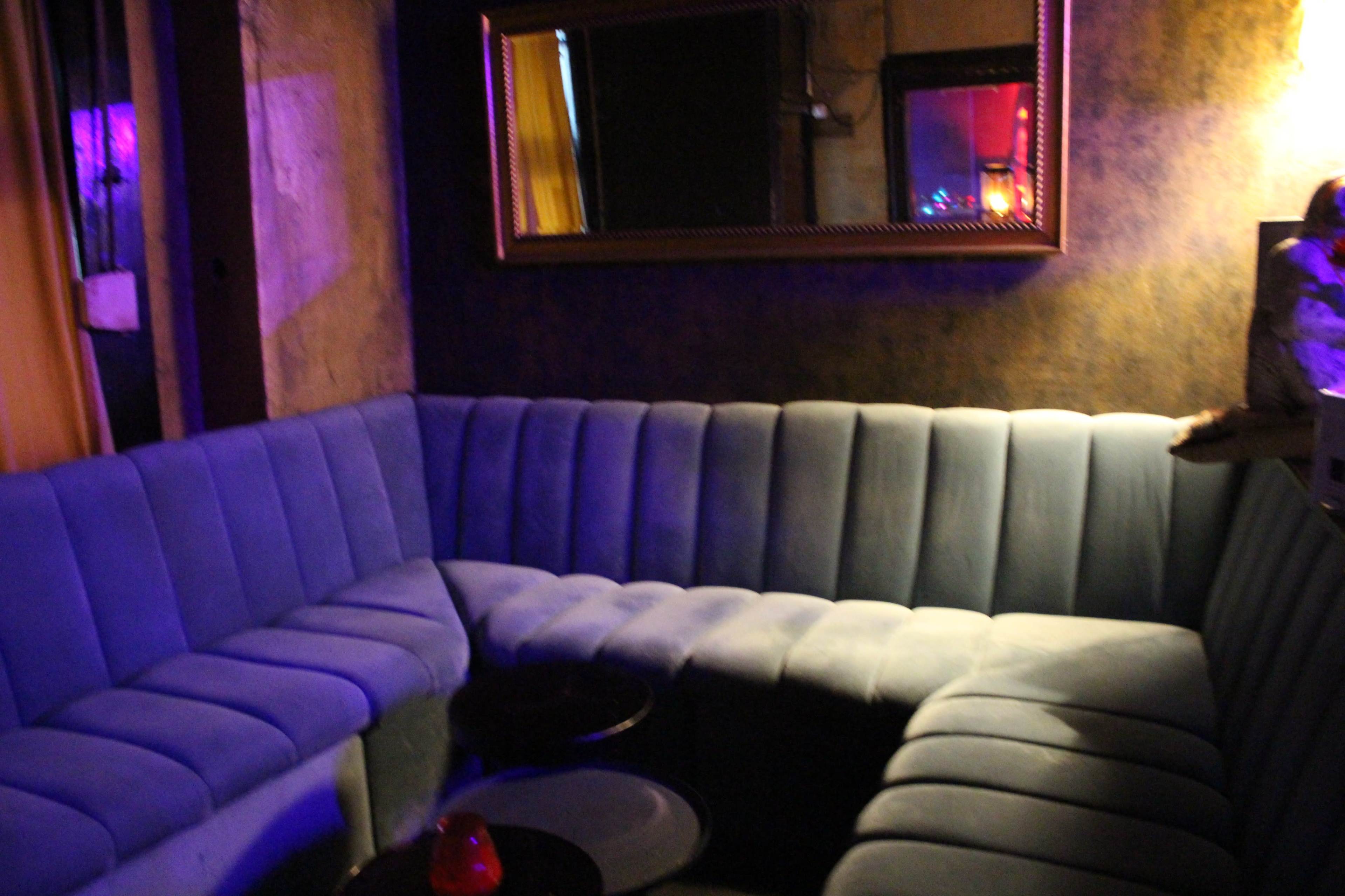 The image shows a dimly lit lounge area with a curved blue upholstery sofa and small round tables.