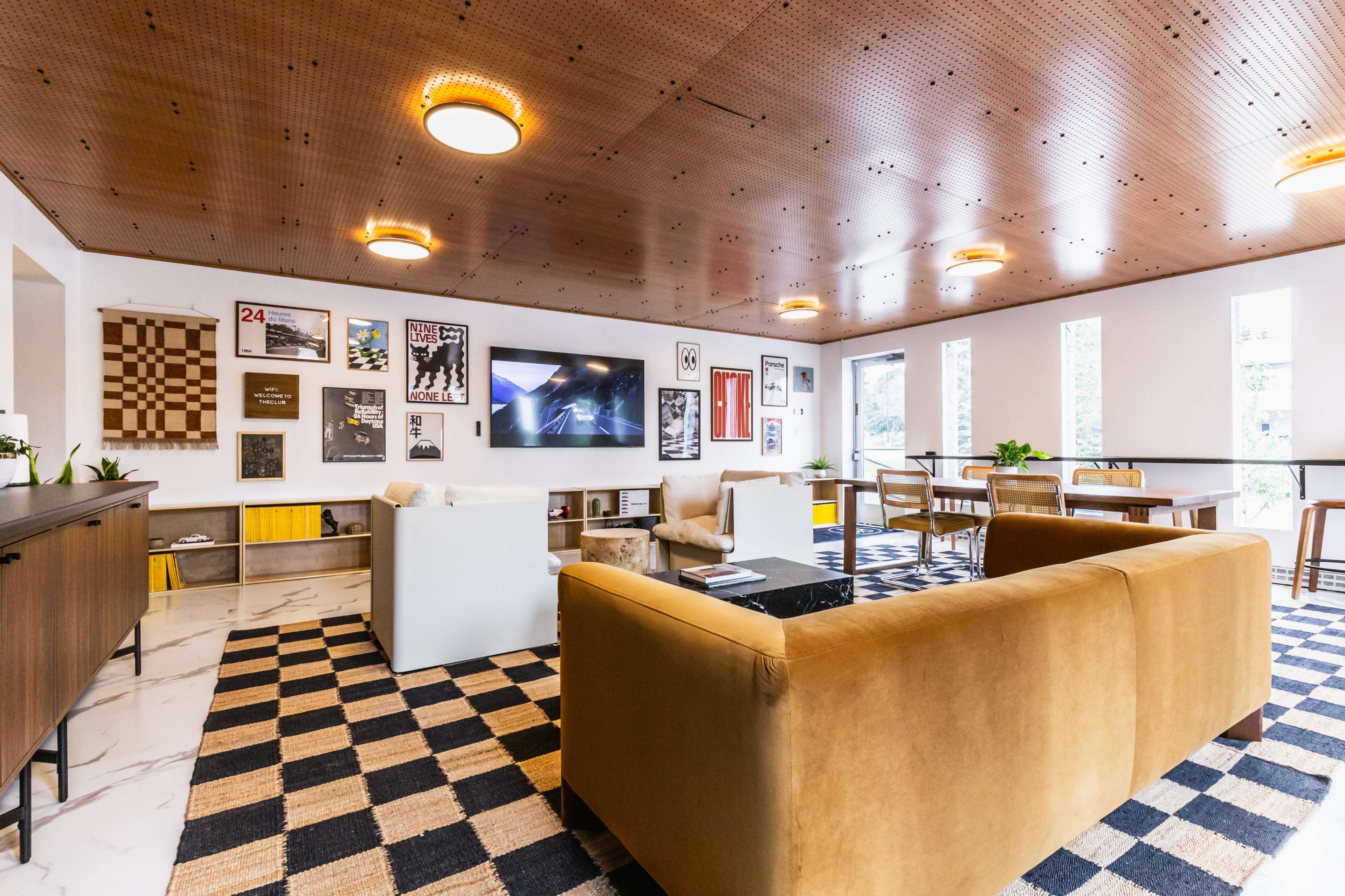 The image shows a modern communal lounge area with a checkered rug, a brown sofa, a television mounted on the wall, and various framed artworks displayed on the walls.