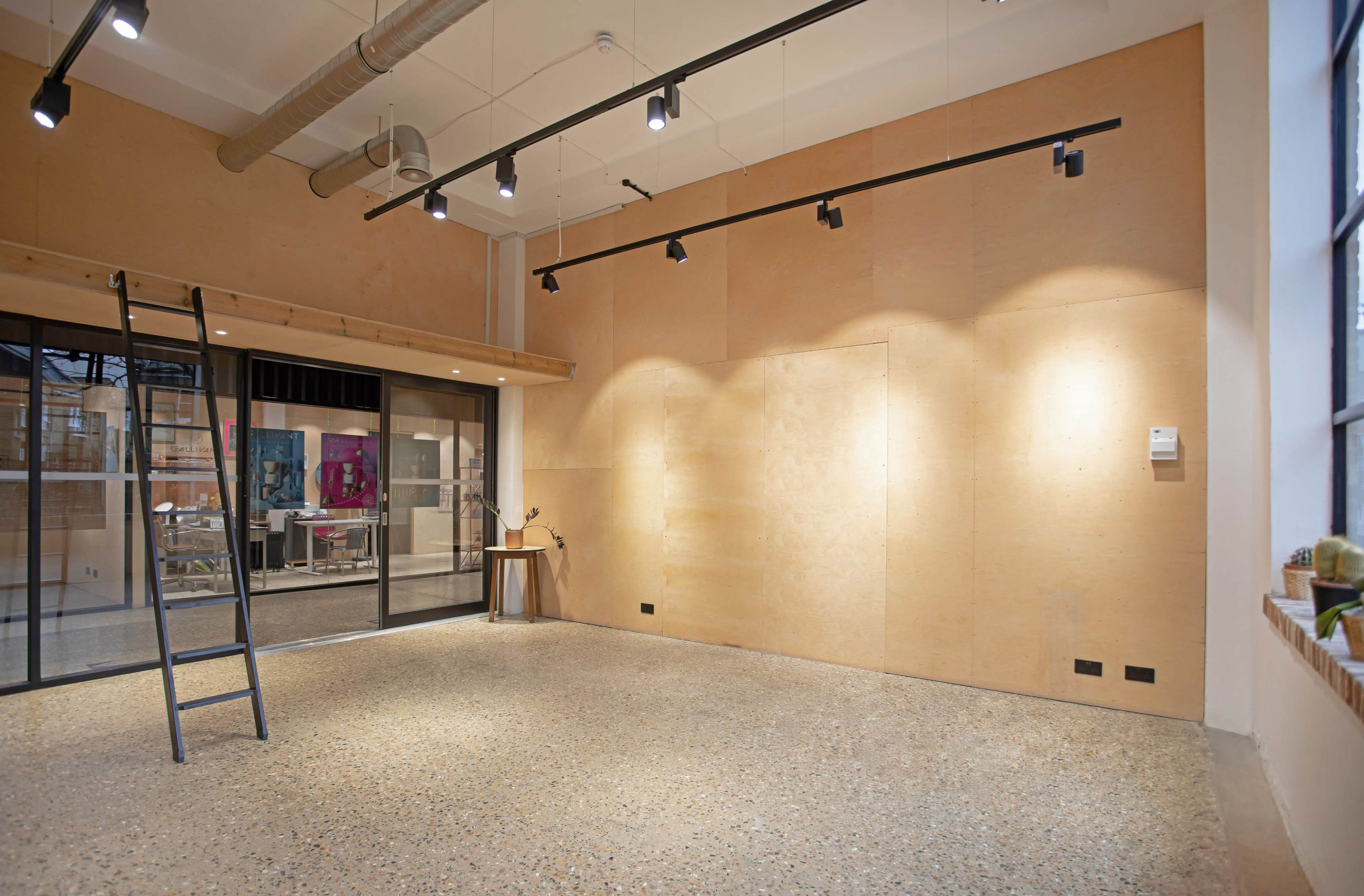 A spacious room with wooden paneling, track lighting, and a concrete floor, featuring a ladder against one wall and glass doors leading to another area.