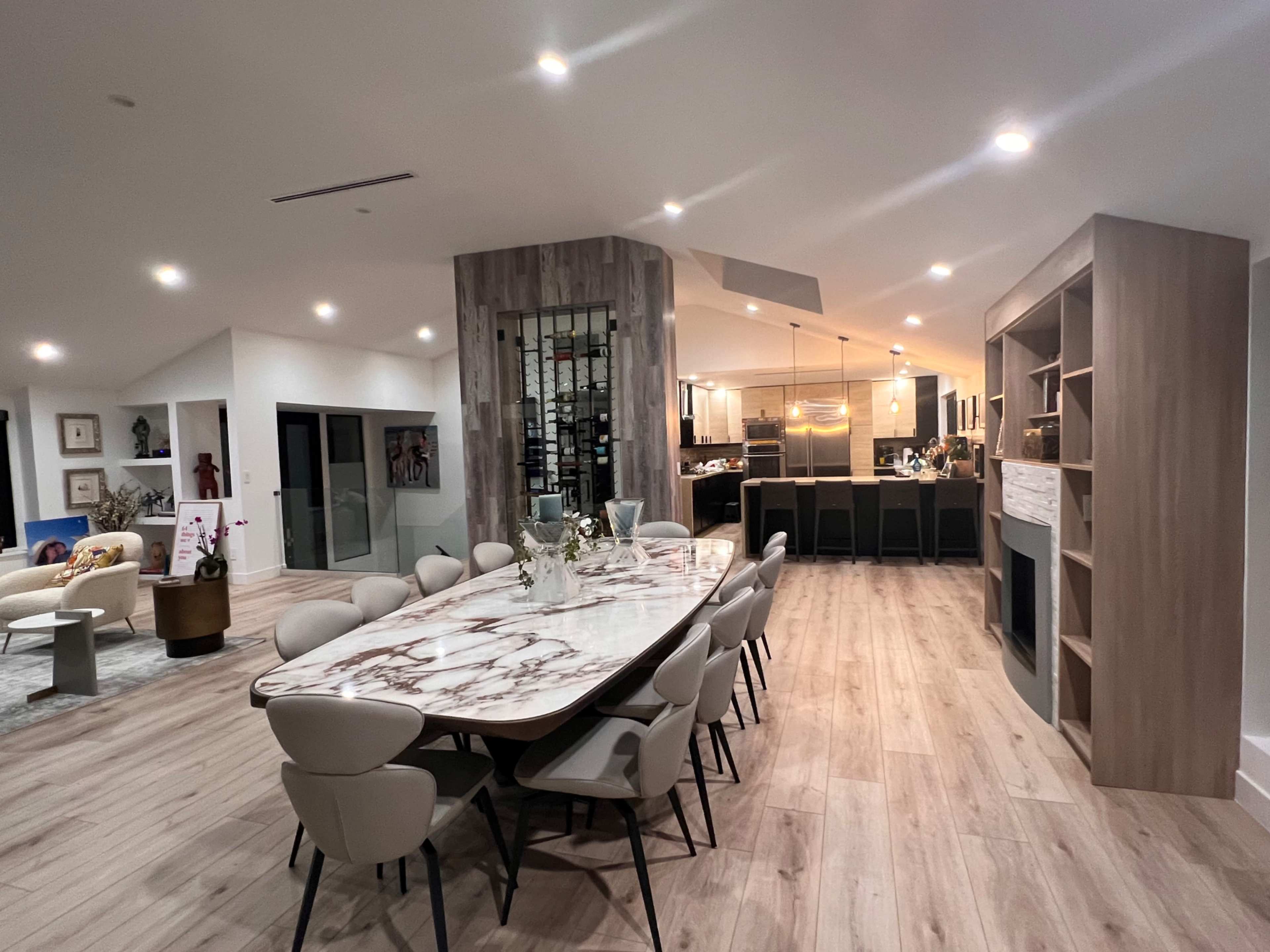 The image shows a modern dining area with a large marble table surrounded by chairs, leading into a kitchen area with a bar counter and various lighting fixtures.