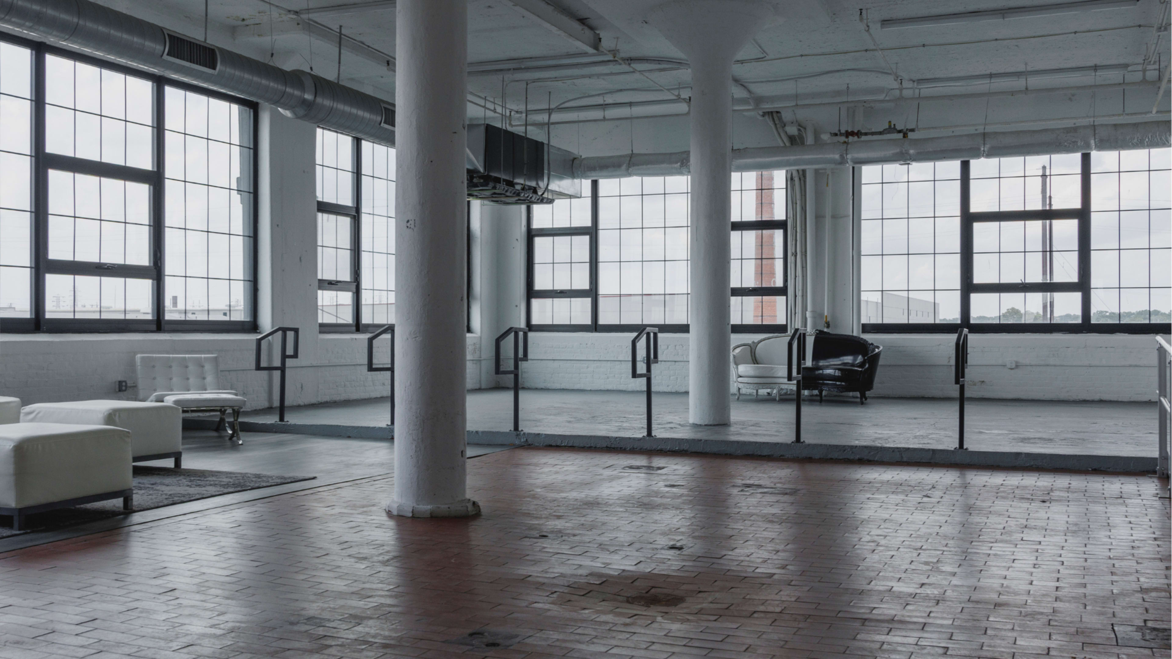 The image shows a spacious industrial-style room with large windows, exposed pipes, and minimal furniture, including a black chair and white seating.