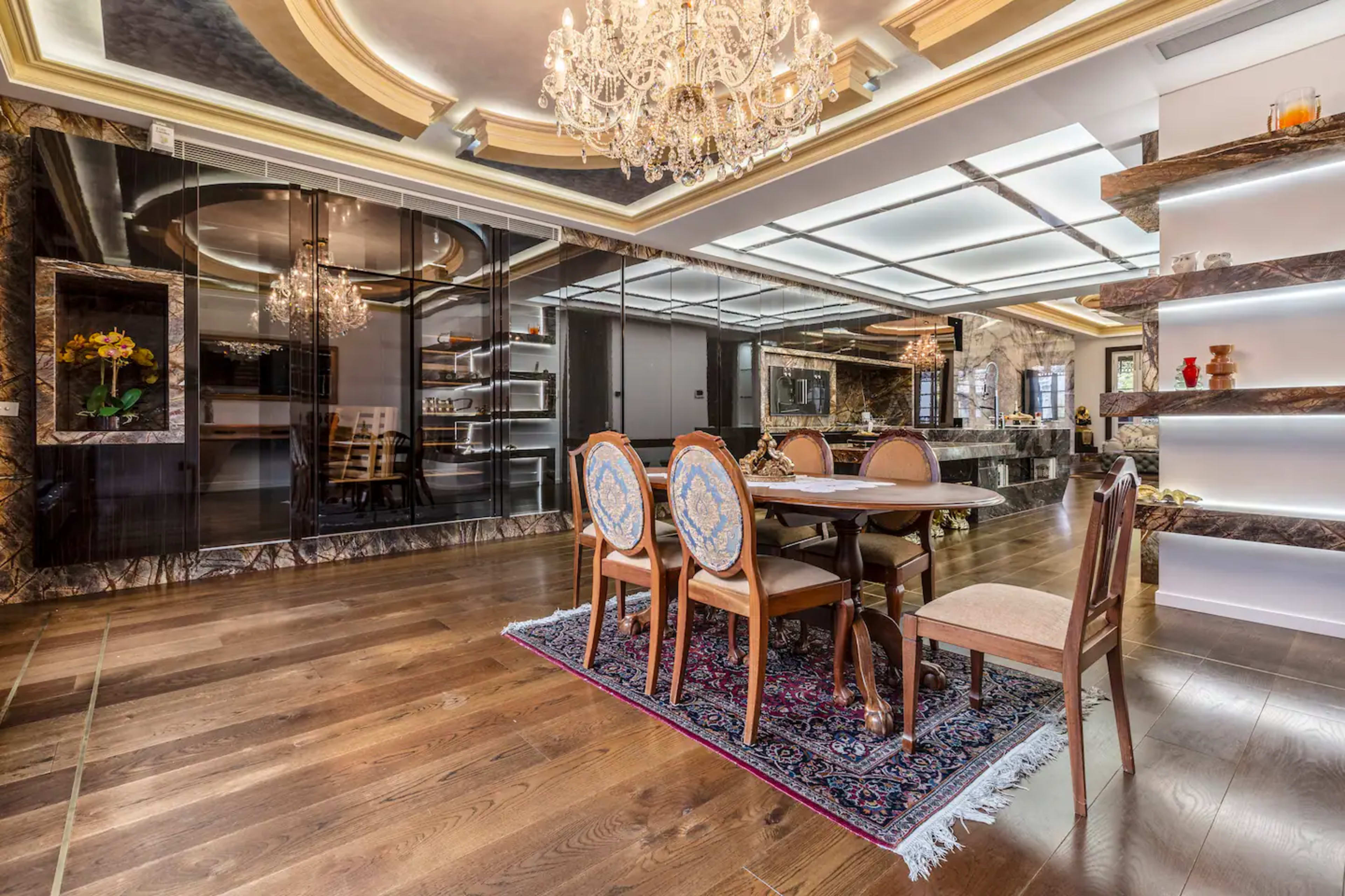 A luxurious dining area with a large table surrounded by elegant chairs, a chandelier overhead, and a marble-finished kitchen in the background.