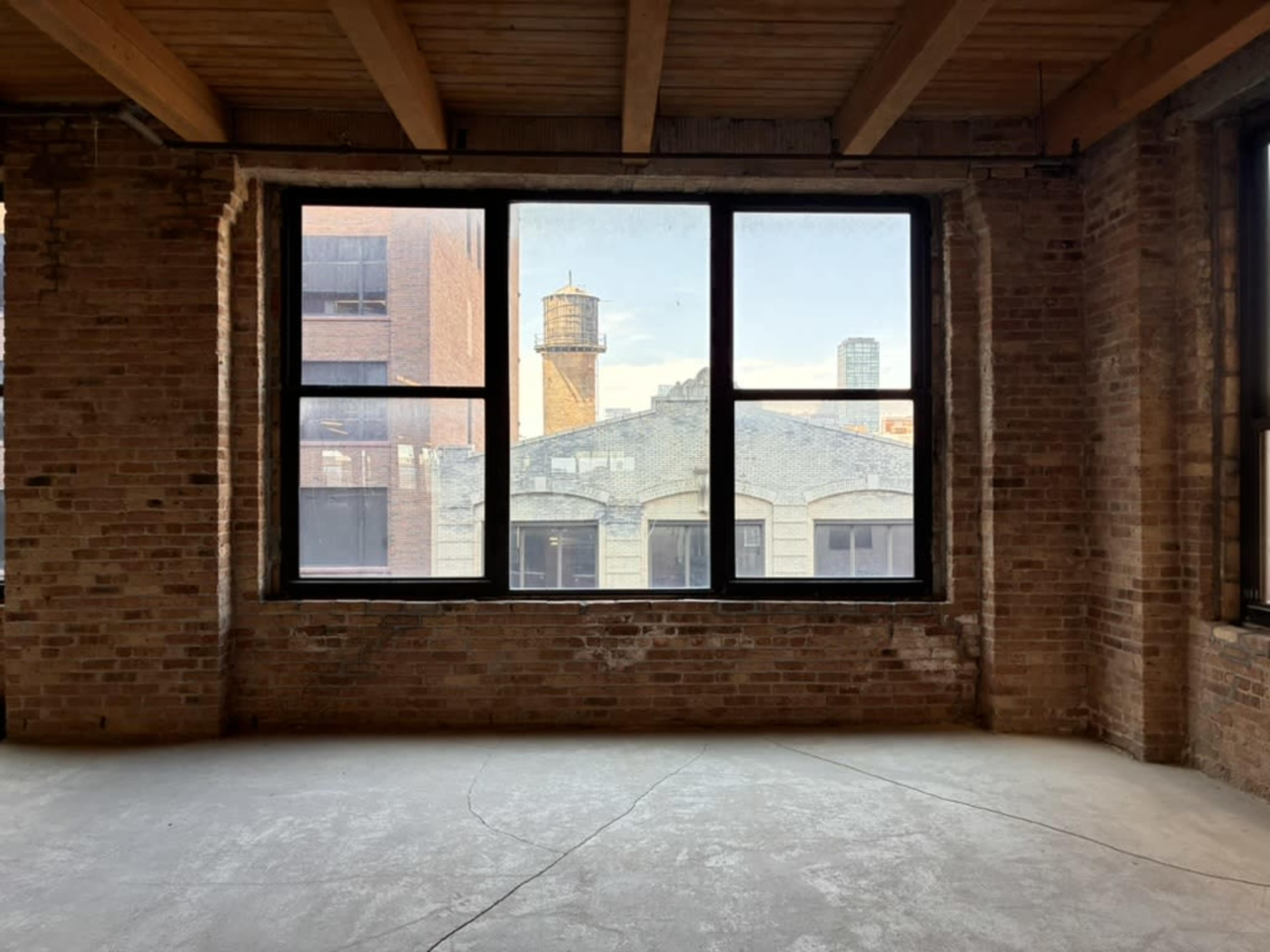 The image shows an empty room with exposed brick walls and large windows overlooking an urban landscape.