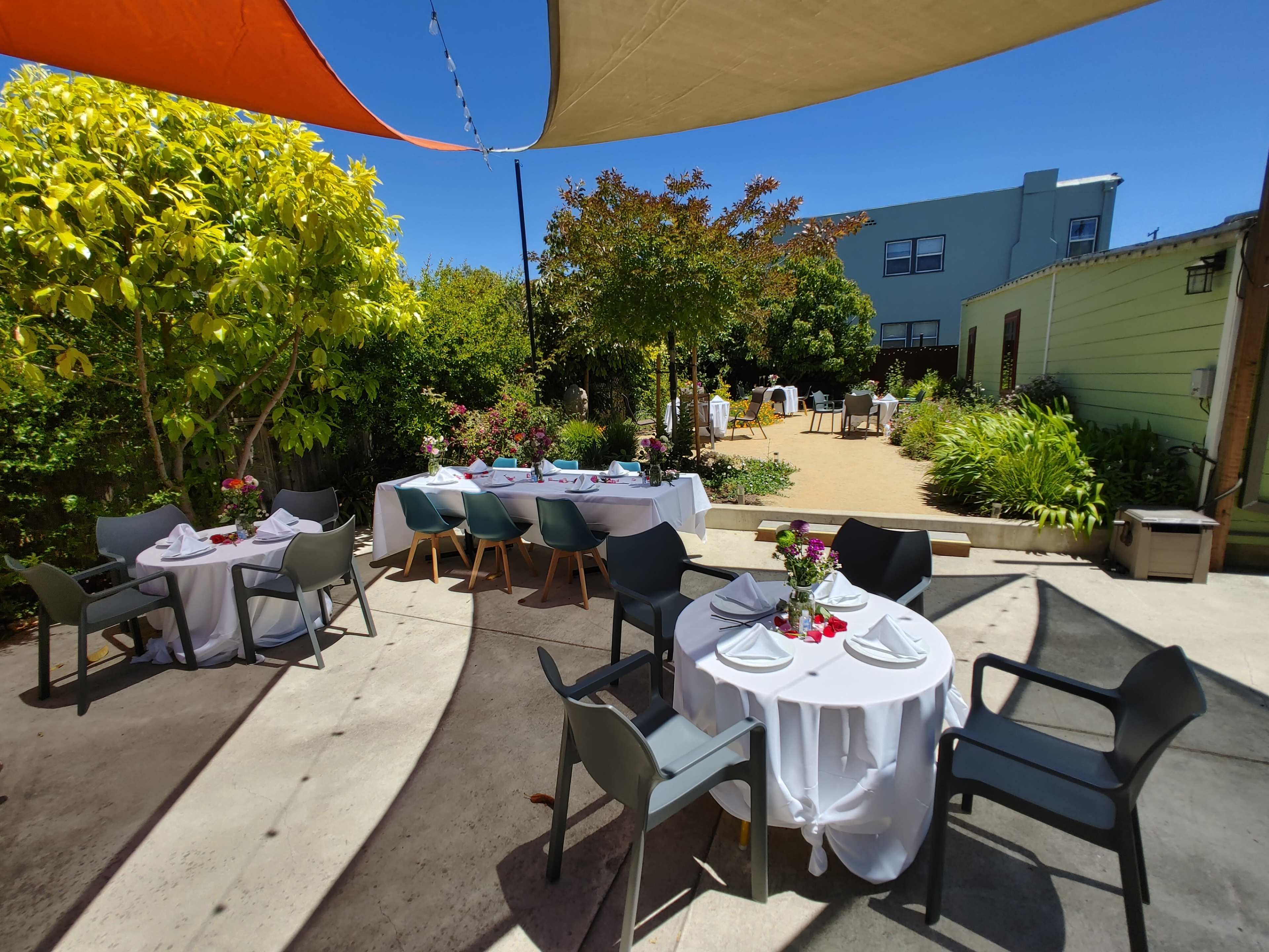 A sunny outdoor dining area features tables set with white tablecloths and flowers, surrounded by greenery and a building in the background.