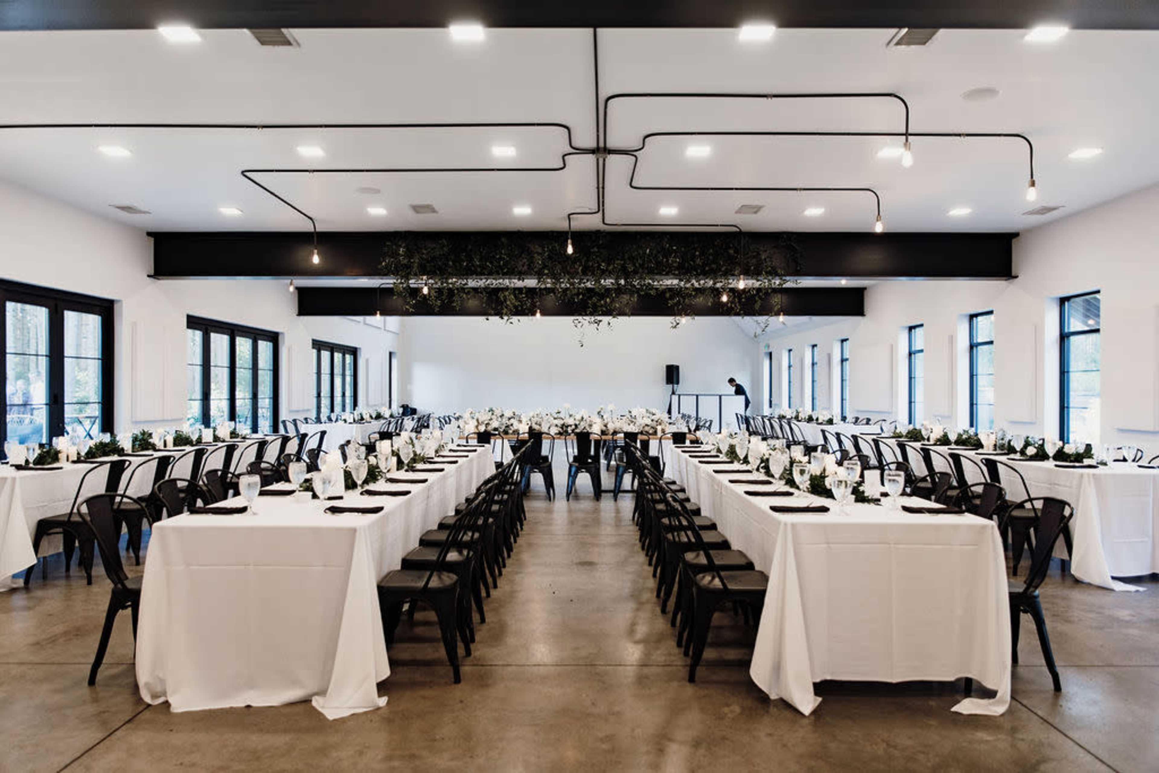 A banquet room with neatly arranged tables covered in white tablecloths, set for an event with centerpieces and chairs facing a stage area.