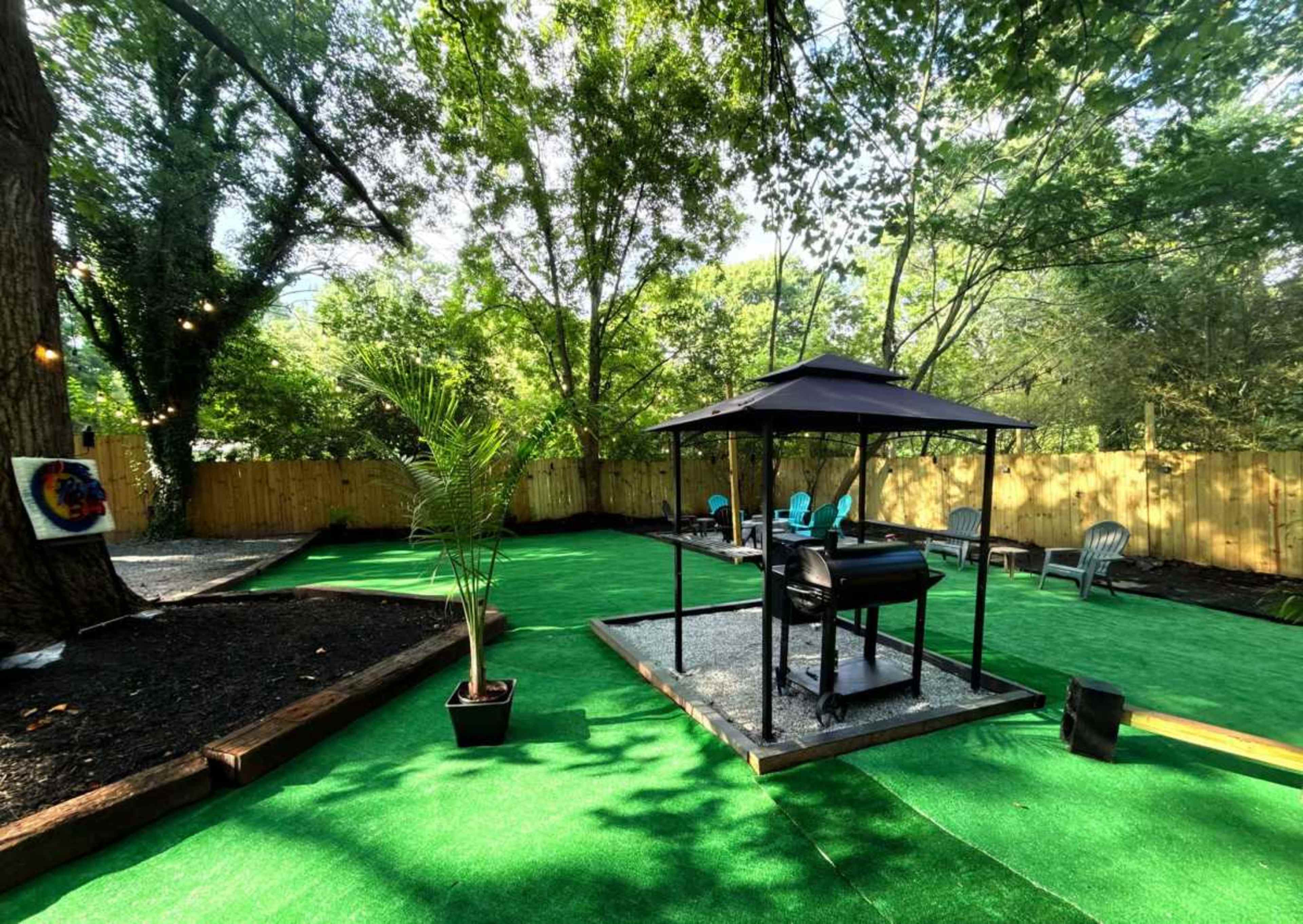 The image shows a backyard with artificial green grass, a gazebo containing a grill, and several outdoor seating areas surrounded by trees and a wooden fence.