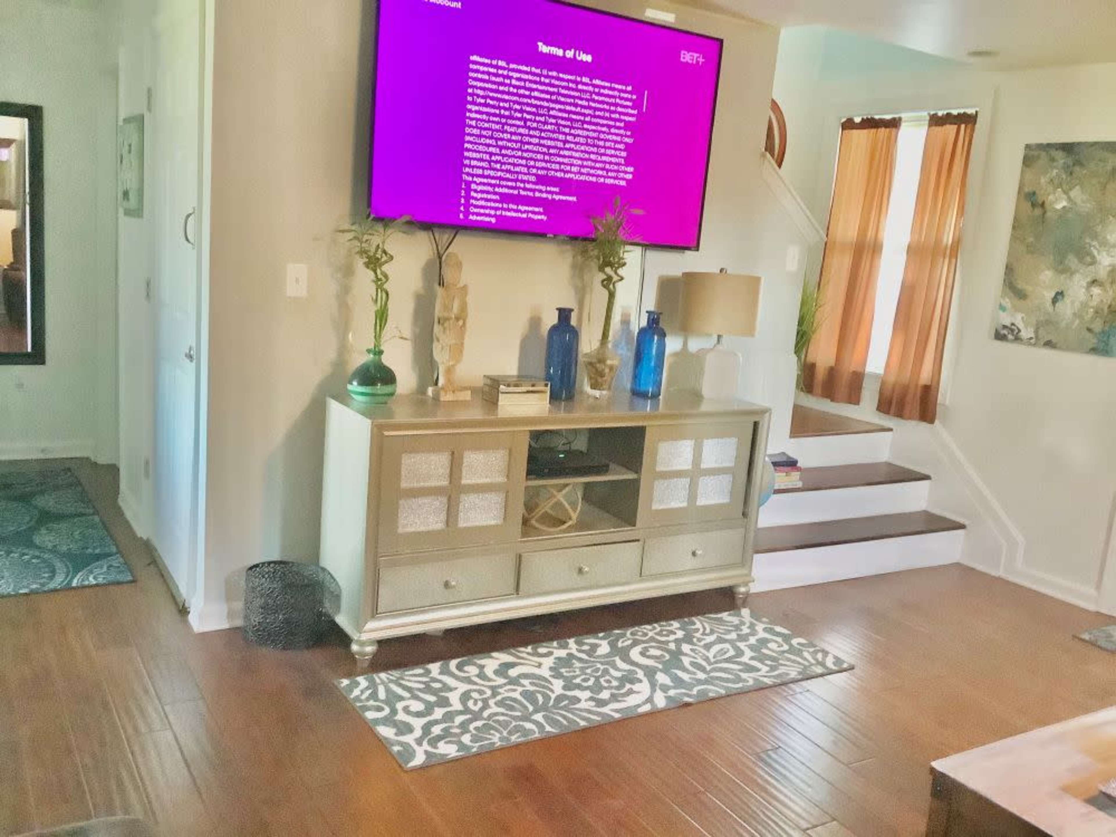 A living room features a mounted TV, a decorative cabinet with vases, and stairs leading to an upper level.