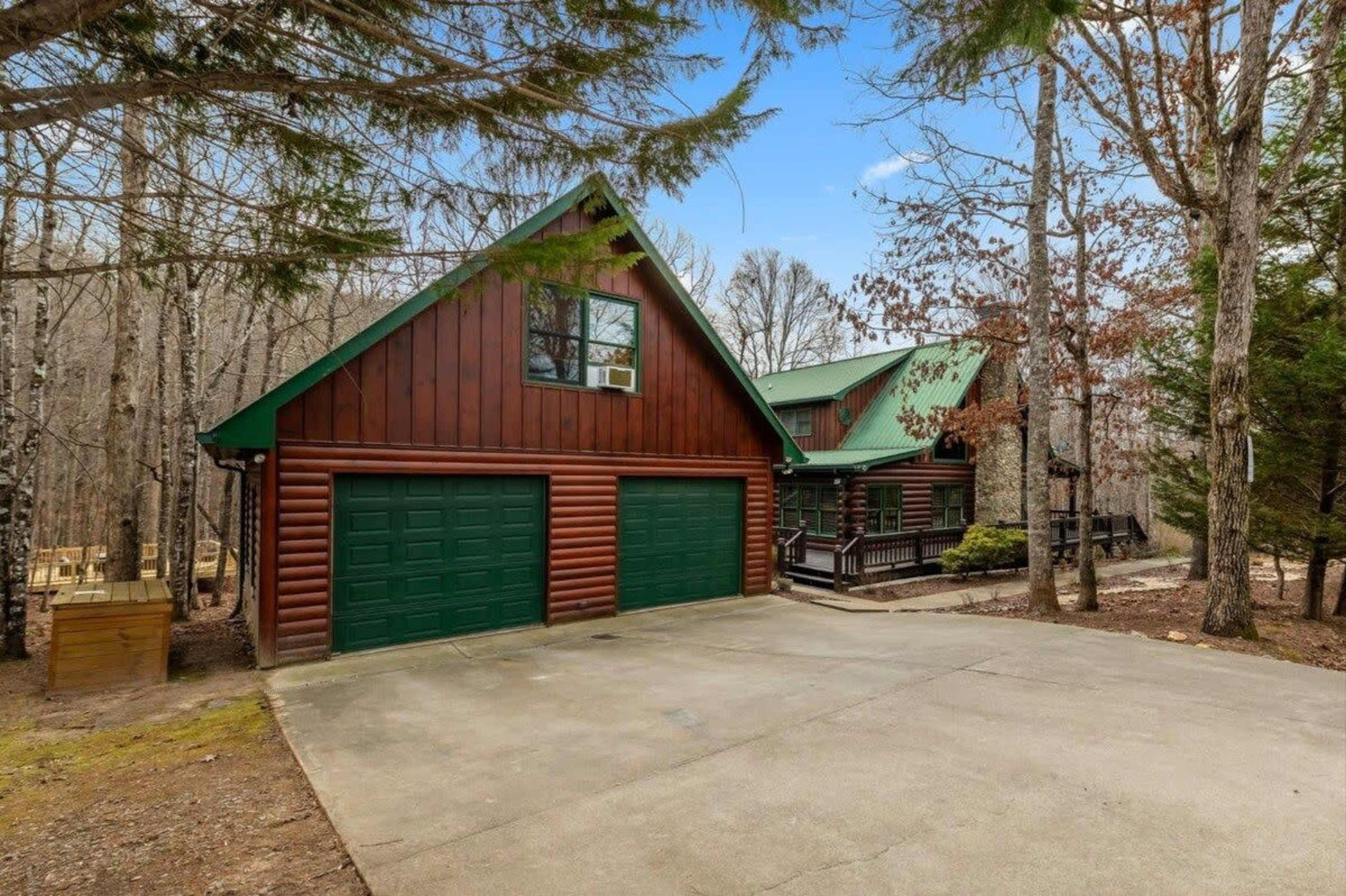 A two-story log cabin with green accents is situated in a wooded area, featuring a concrete driveway and garage doors.