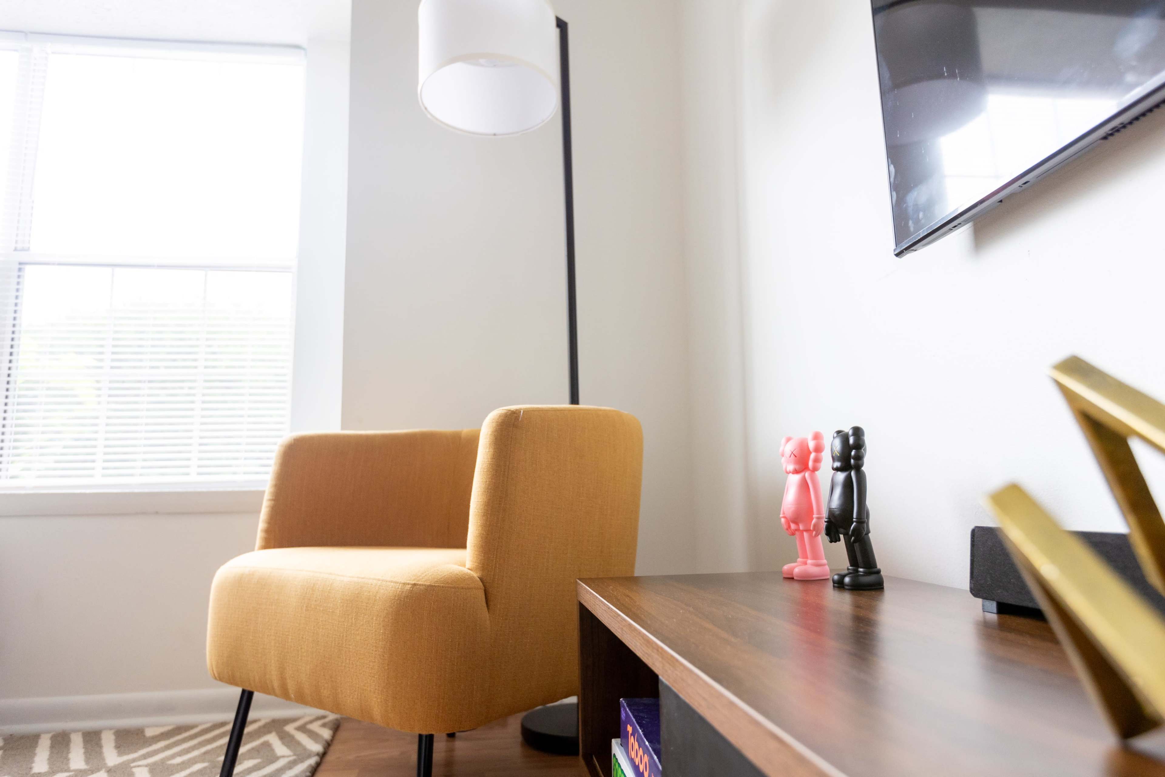A cozy corner of a living room features an orange chair beside a floor lamp, with a wooden shelf displaying colorful figurines and a television mounted on the wall.
