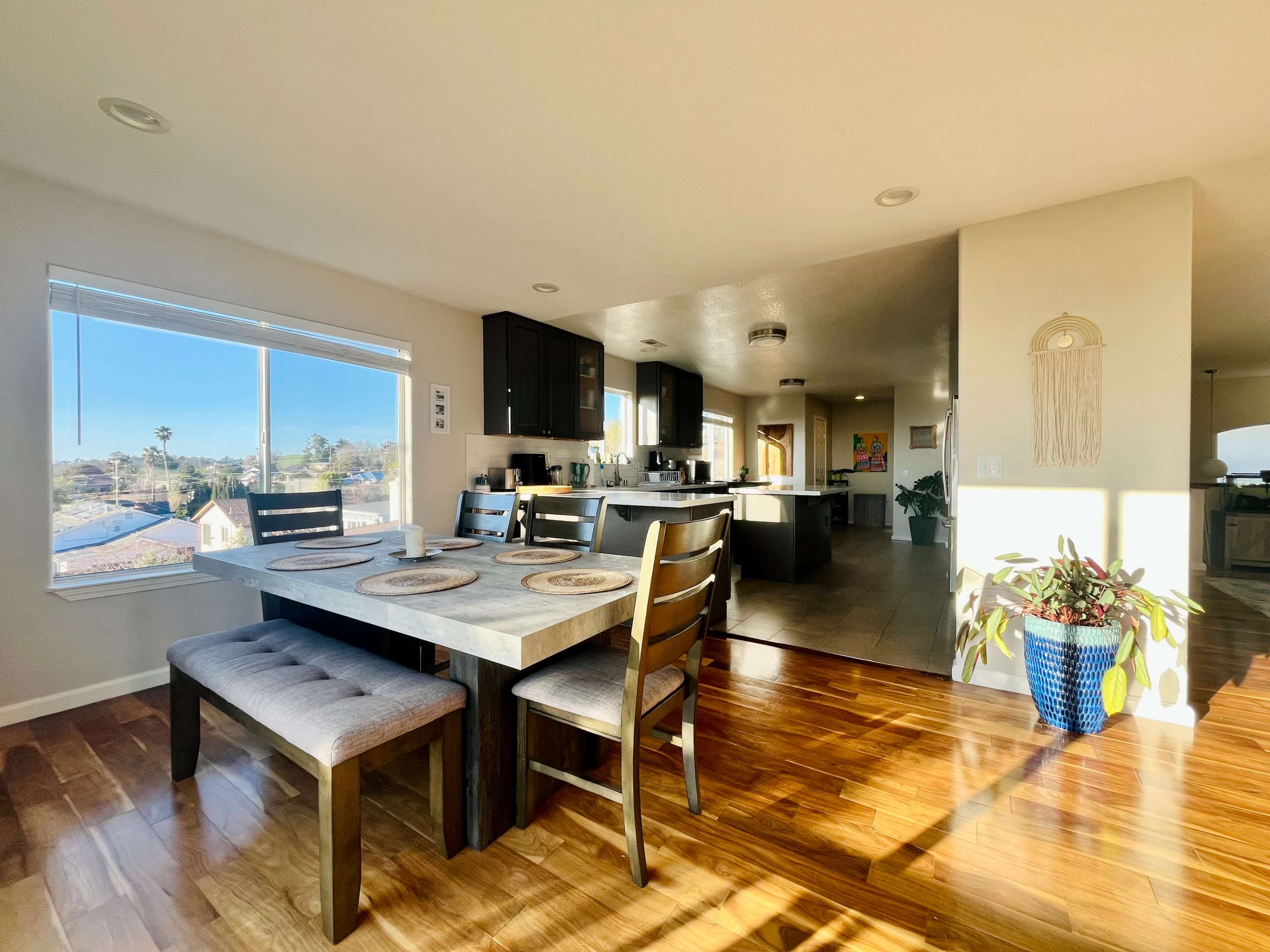 A modern kitchen and dining area featuring a rectangular table with chairs, large windows allowing natural light, and dark cabinetry.