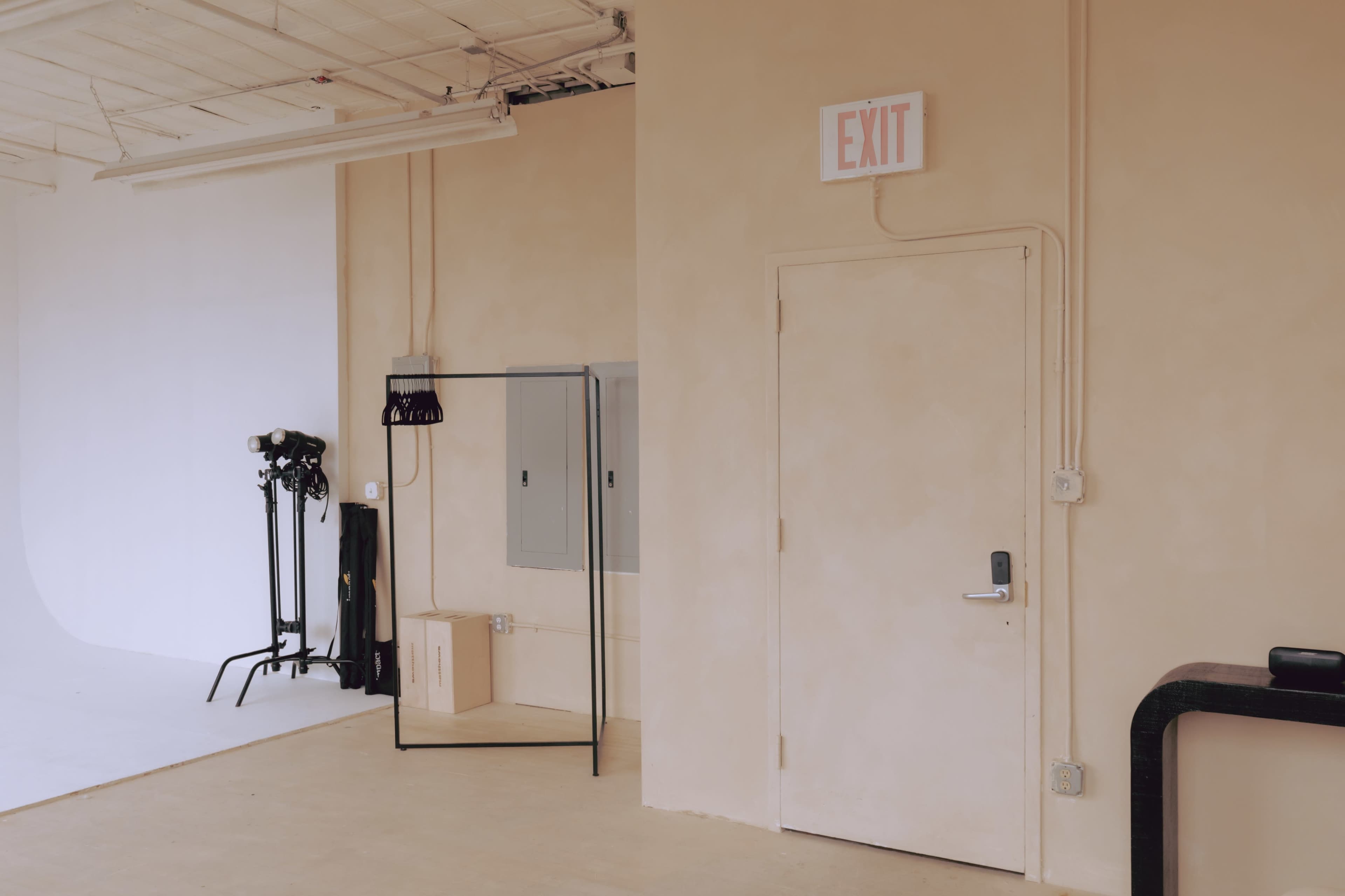 A minimalistic room features an EXIT sign above a door, a mirror, a gray electrical panel, and photography equipment on the floor.