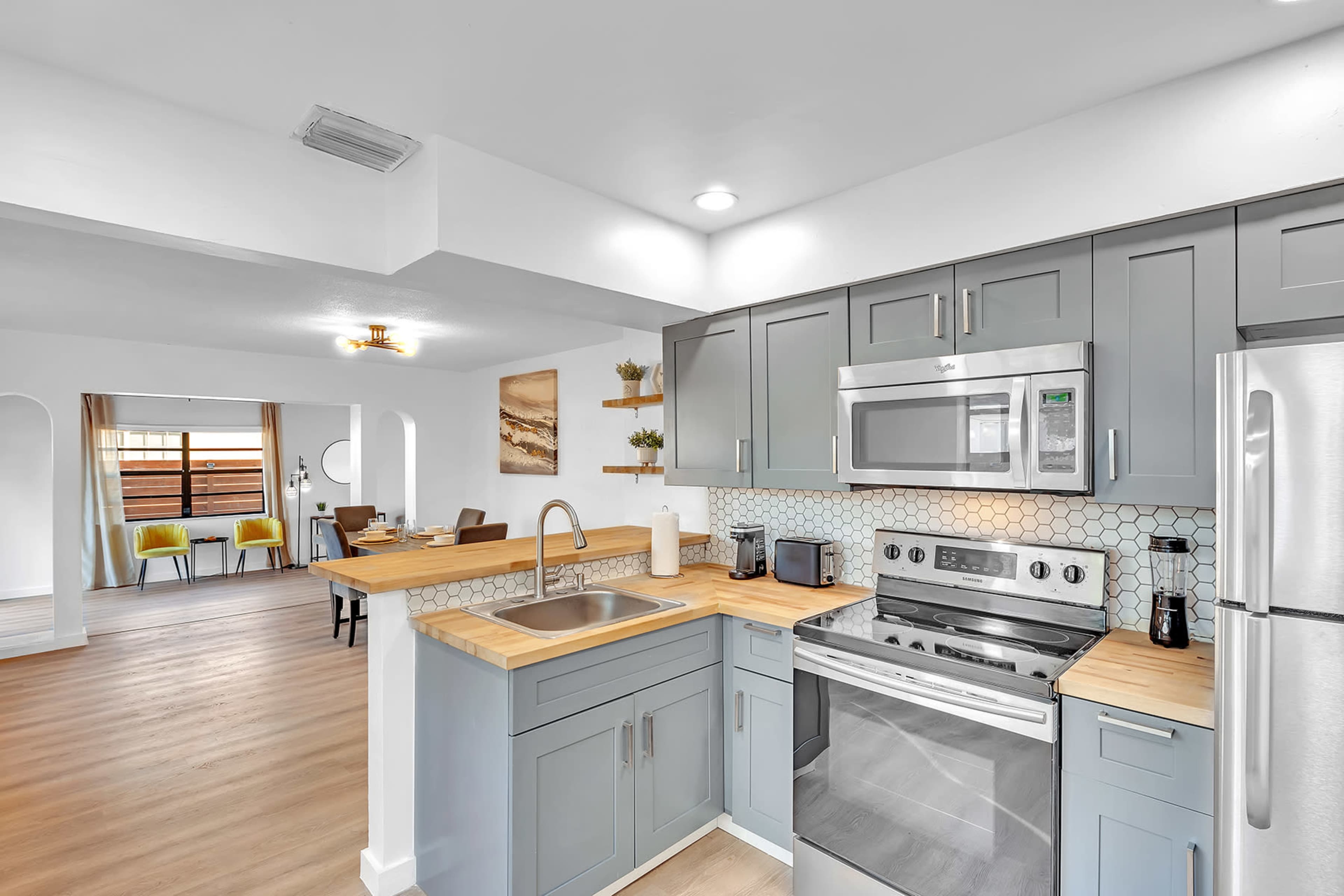A modern kitchen features gray cabinetry, stainless steel appliances, and a wooden countertop, with an open layout leading to a dining area in the background.