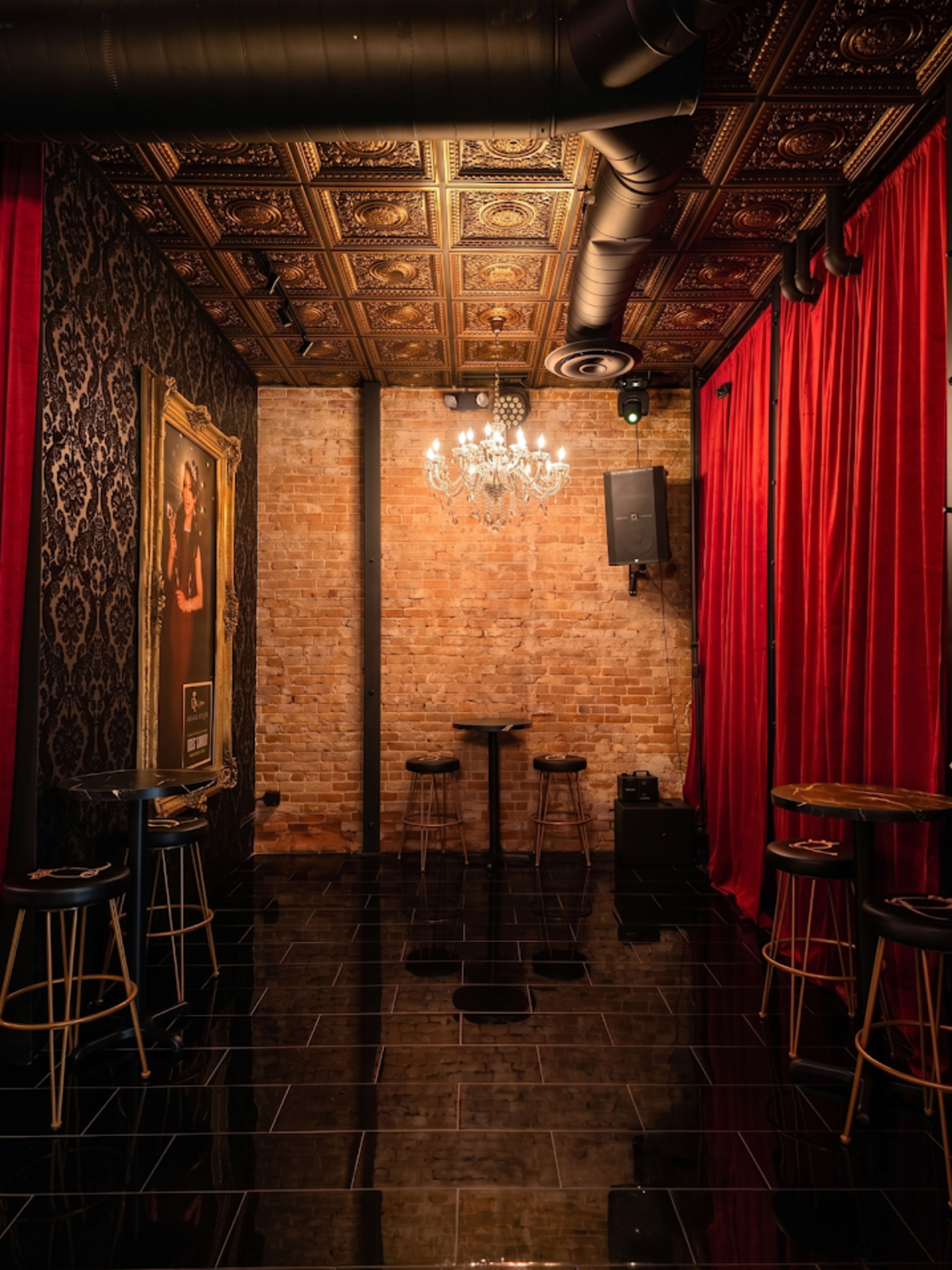 The image shows an elegantly designed room with exposed brick walls, black tiled floors, a chandelier, and red velvet curtains.