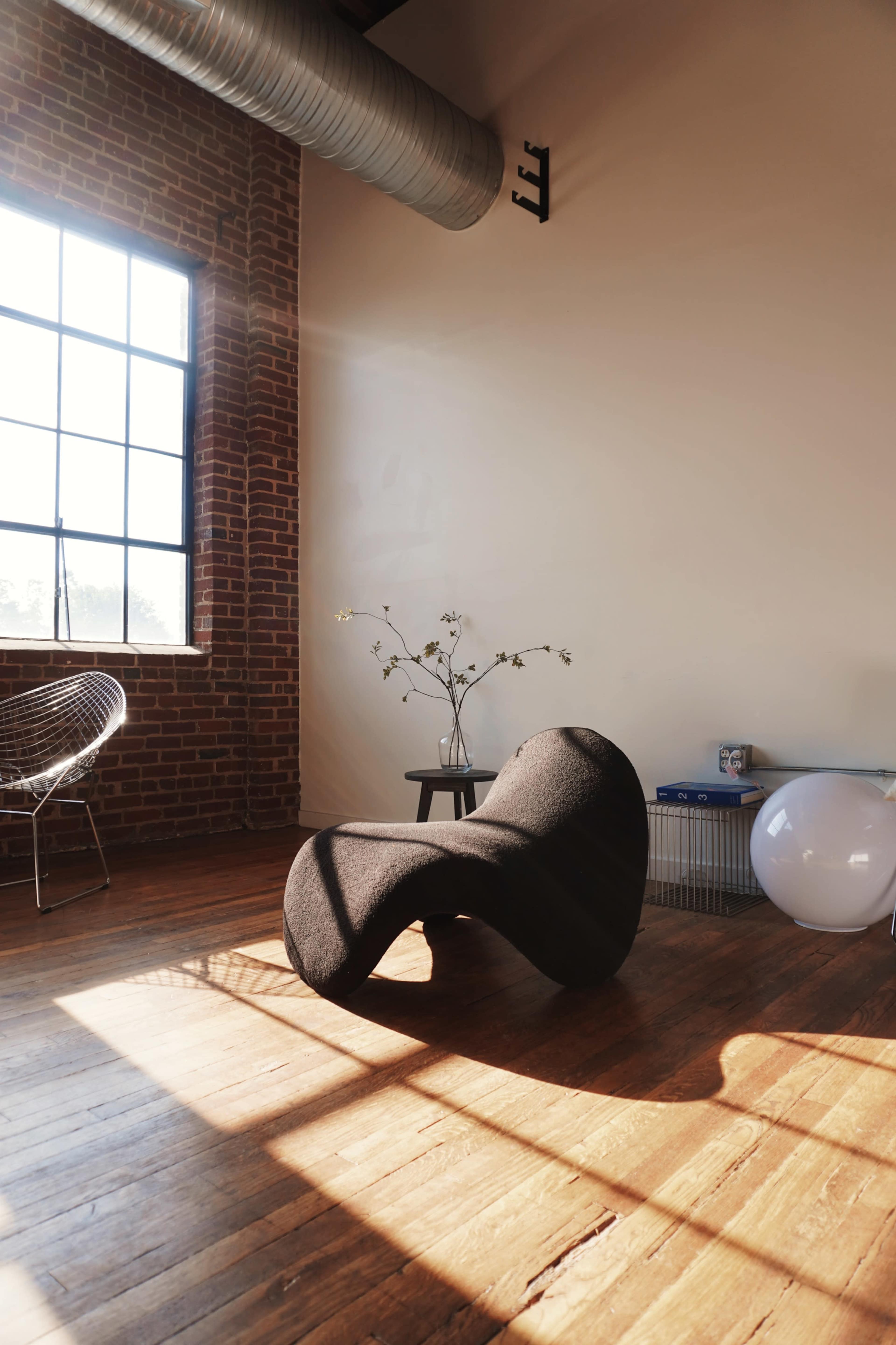 A curved black chair is positioned on a wooden floor beside a small table with a vase of flowers, illuminated by sunlight streaming through a large window.