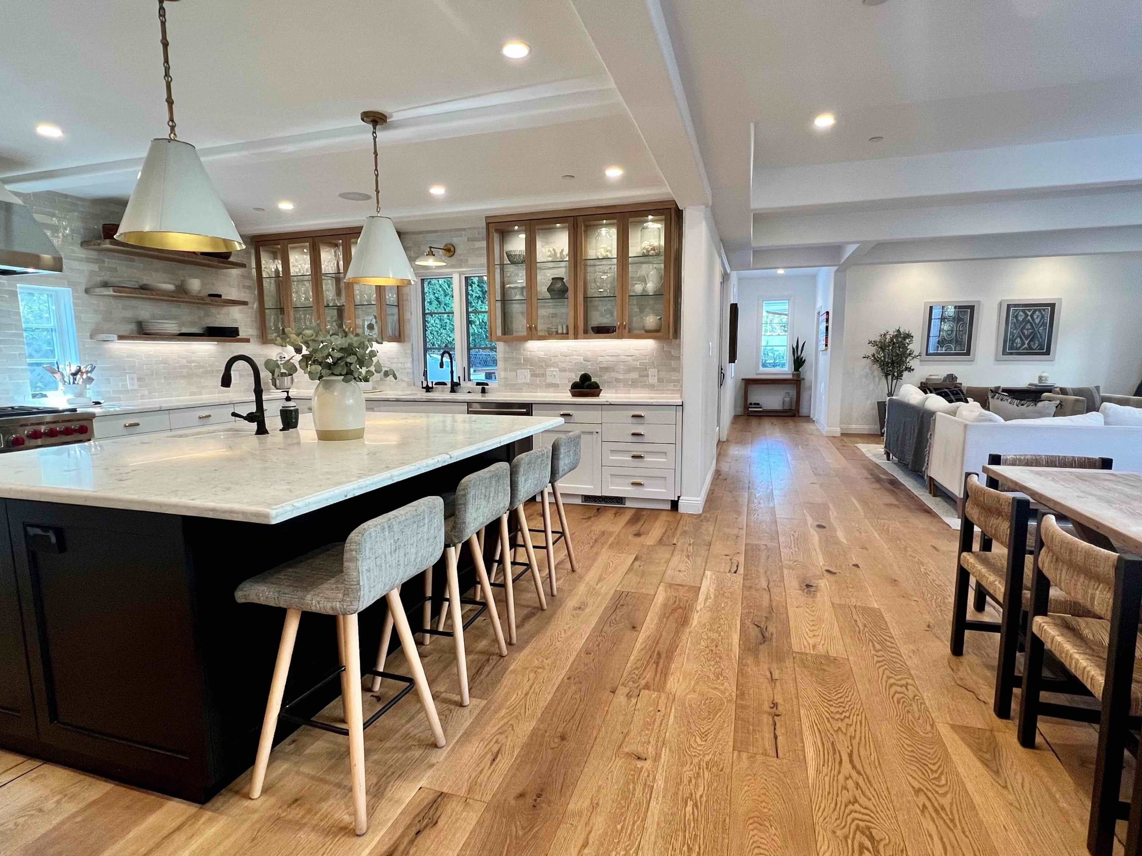 A modern kitchen with a large marble island, wooden flooring, and an open living area in the background.