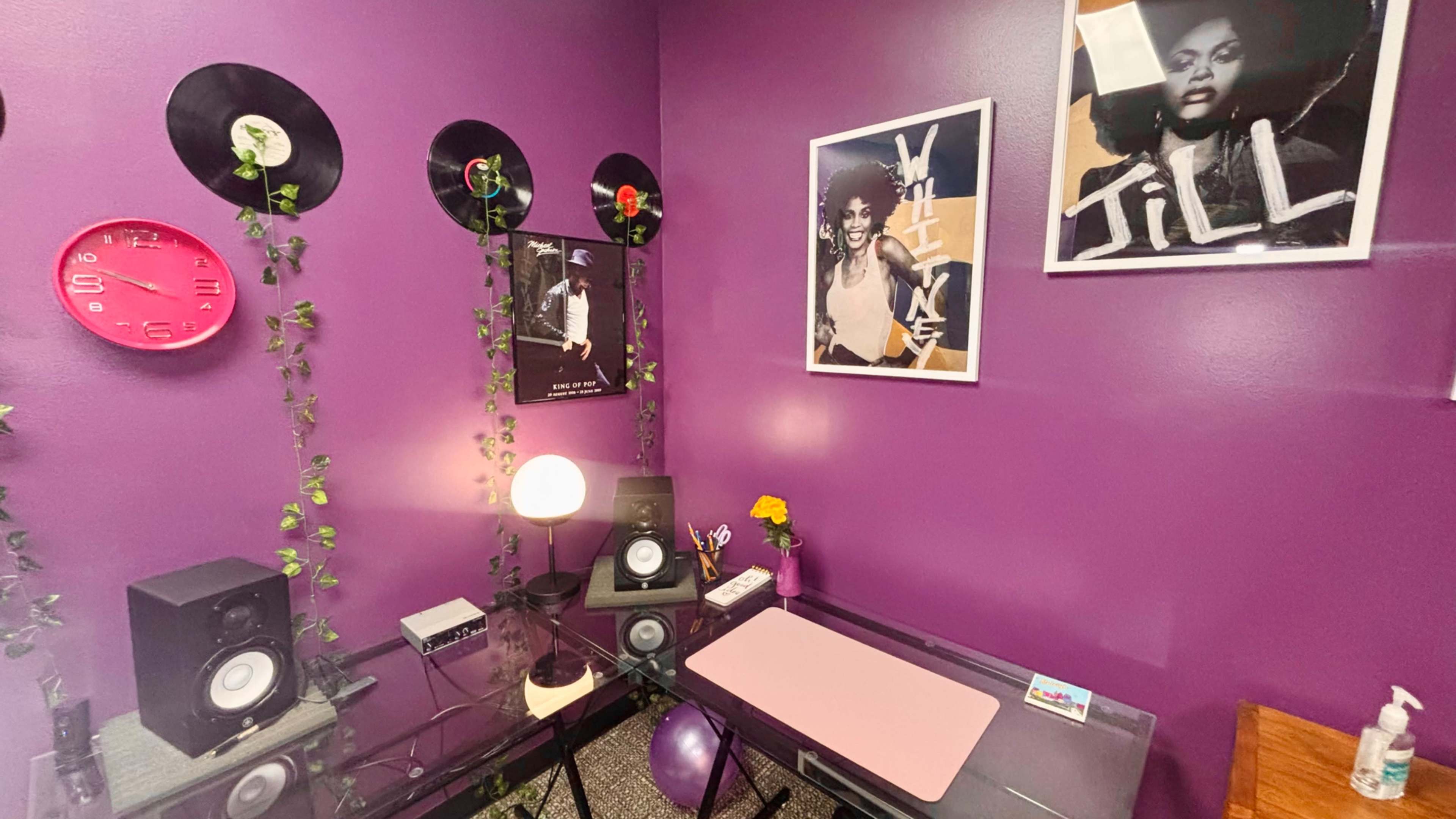 The image shows a small room with purple walls, featuring a glass desk, vinyl records on the walls, and framed photos of musicians alongside a clock and a lamp.