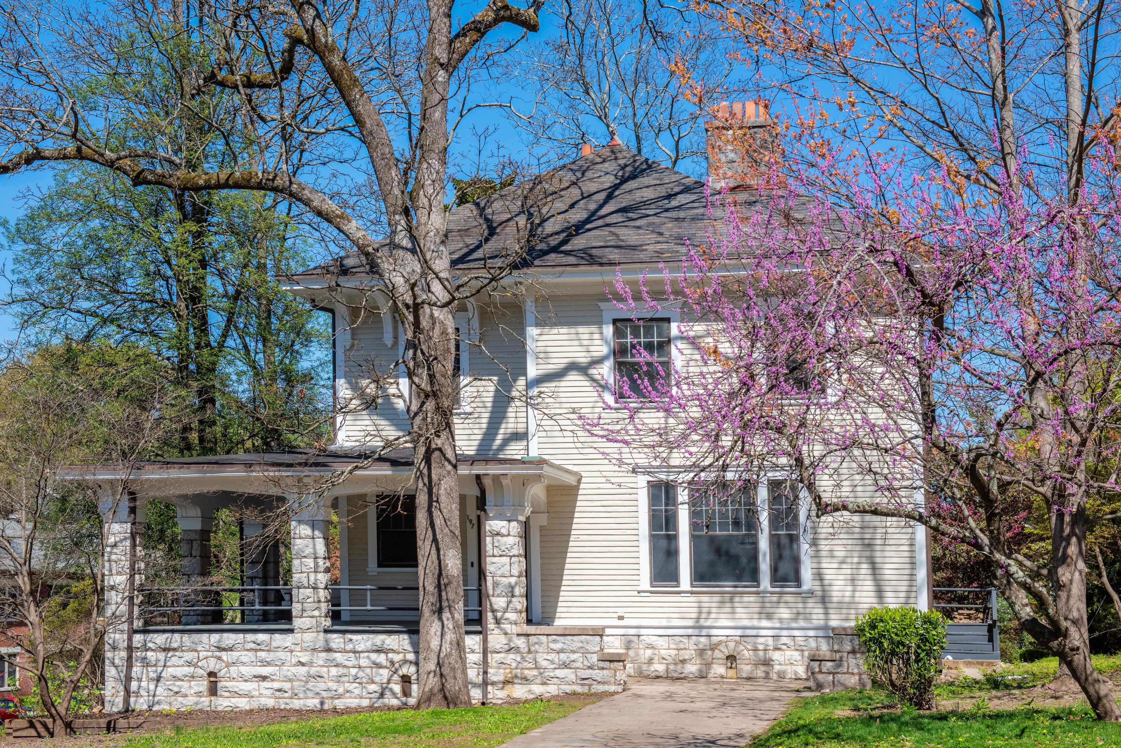 A two-story house with a front porch and stone accents is set against a backdrop of green trees and a blooming pink tree under a clear blue sky.