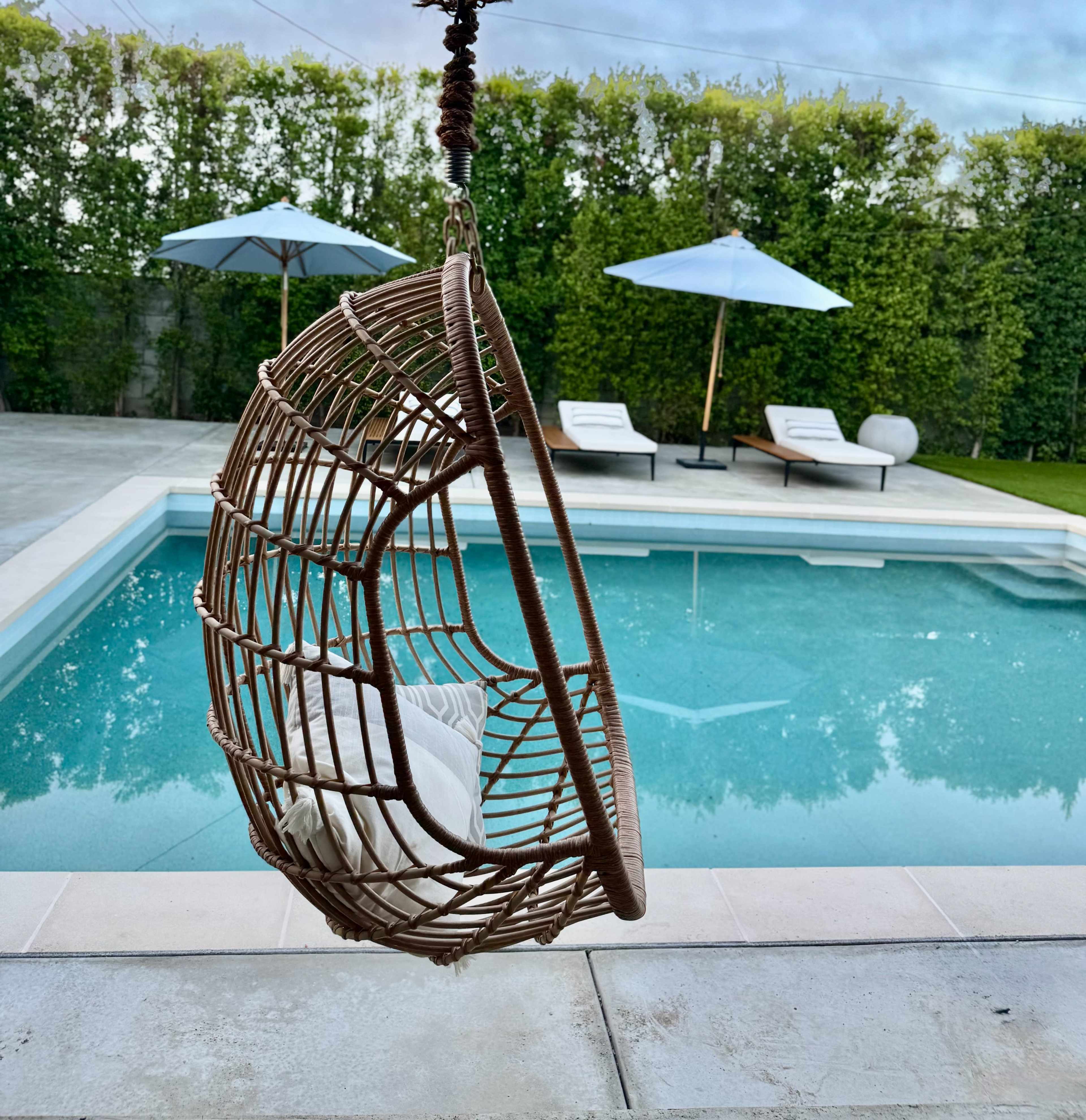 A rattan hanging chair with a cushion is positioned near a swimming pool surrounded by lounge chairs and umbrellas.