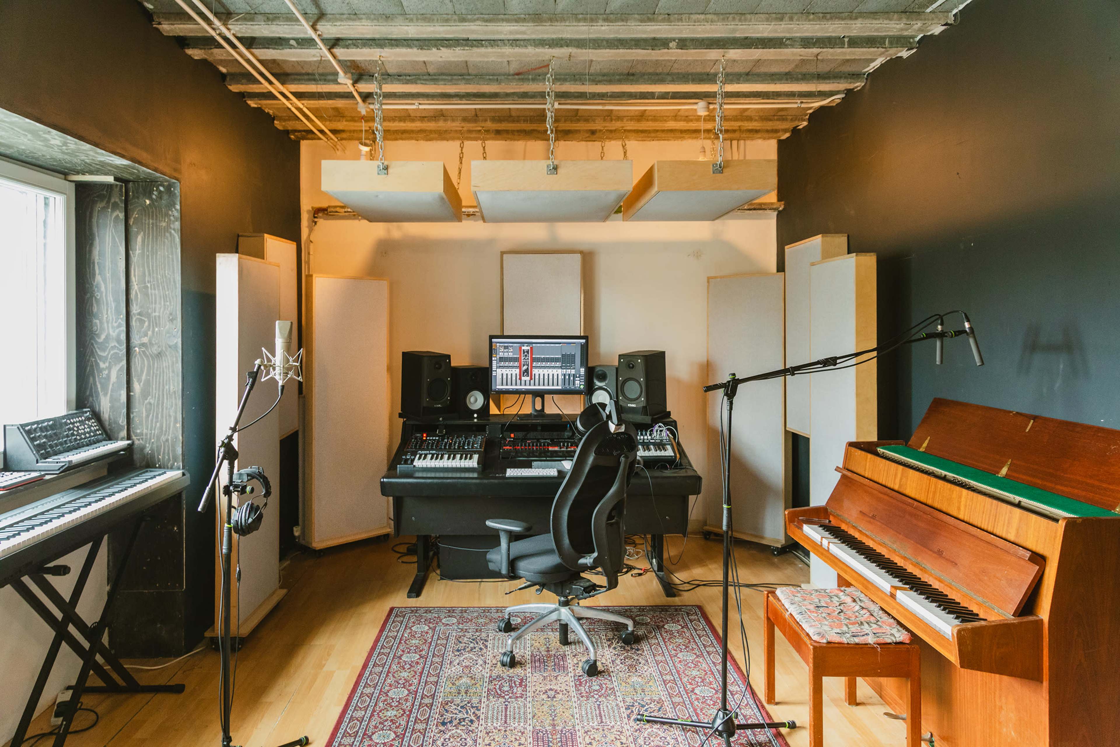 The image depicts a music production studio featuring a central desk with a computer setup, various instruments, soundproof panels, and microphones arranged around the room.