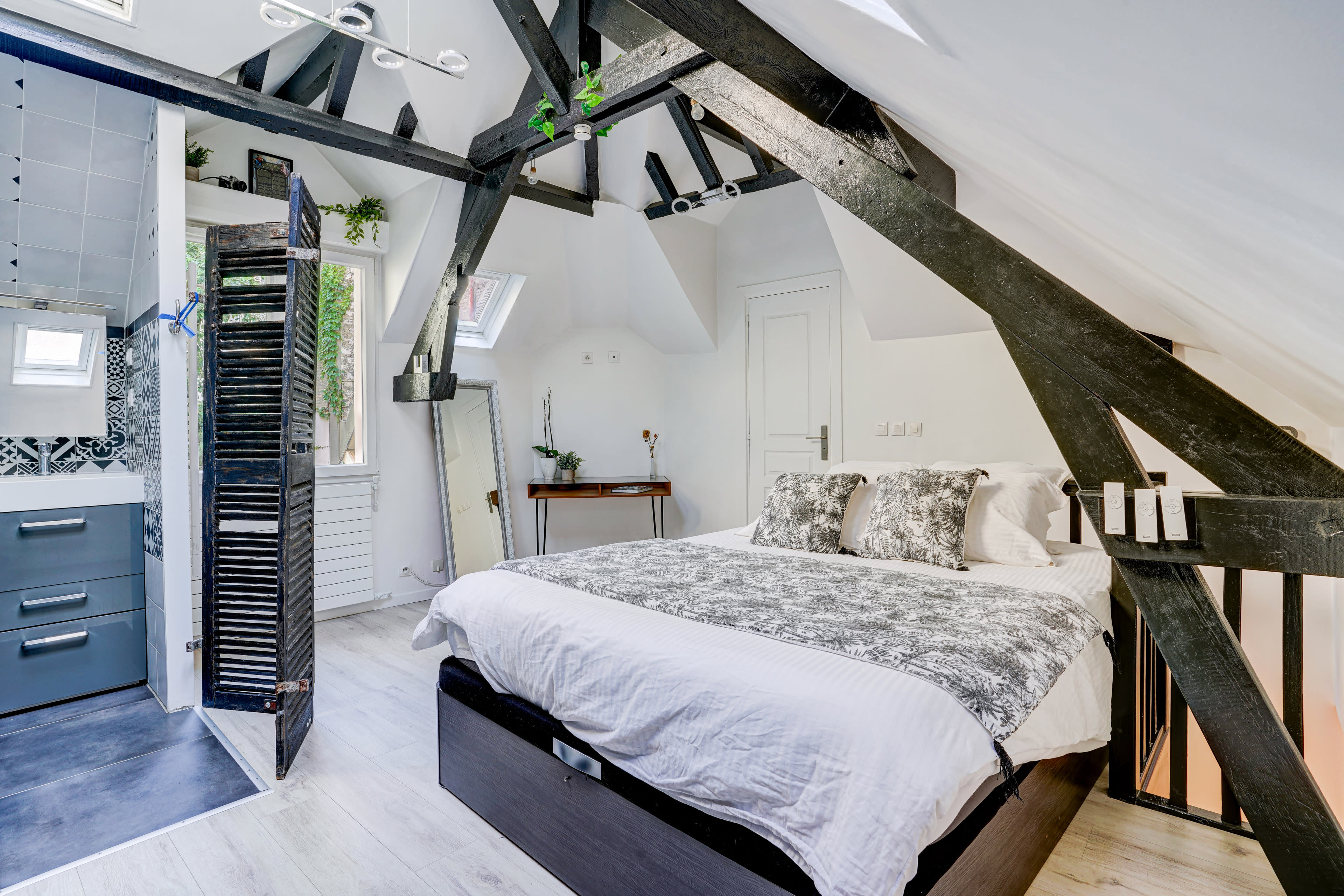 The image shows an attic bedroom featuring a bed with decorative pillows, exposed wooden beams, and an adjoining bathroom area with modern fixtures.