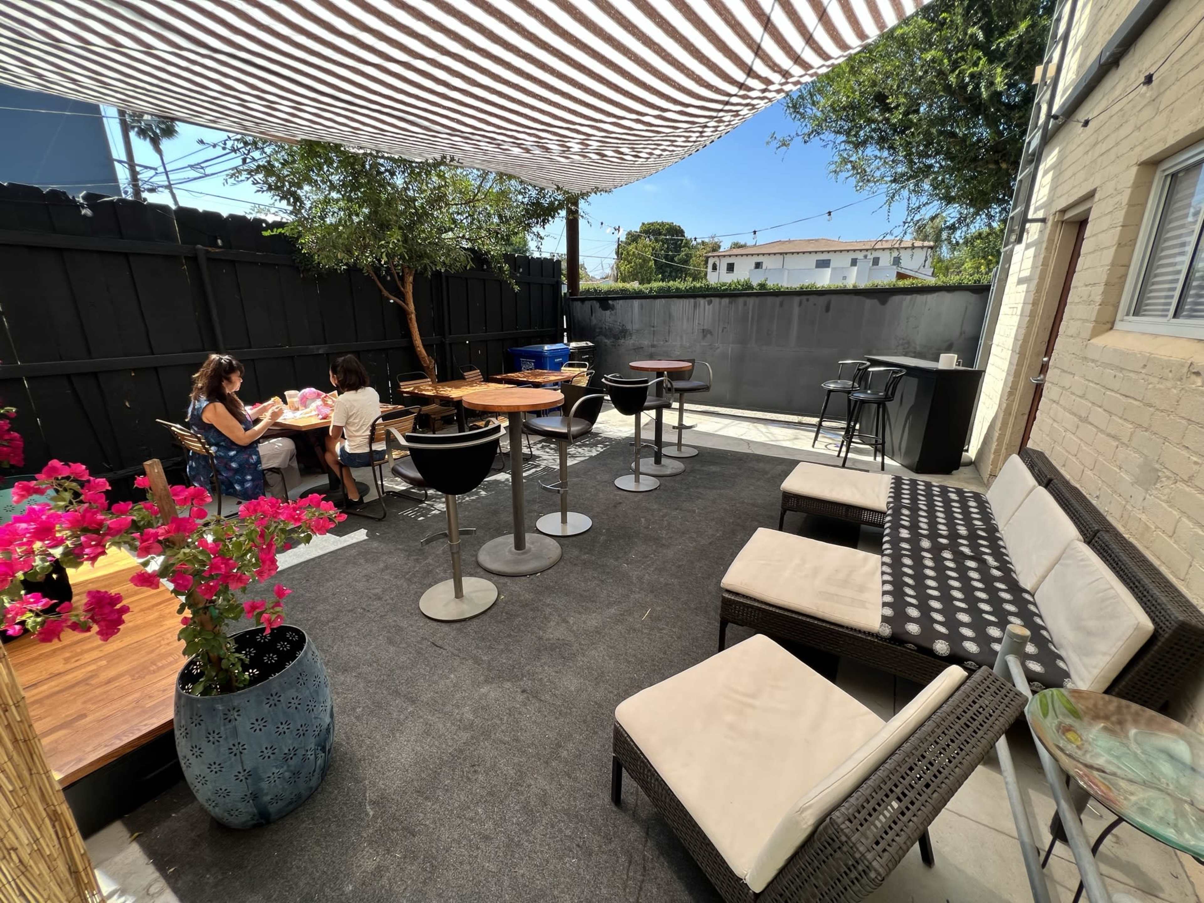 A patio area features tables, chairs, and seating surrounded by plants, with a striped canopy overhead and a black fence in the background.