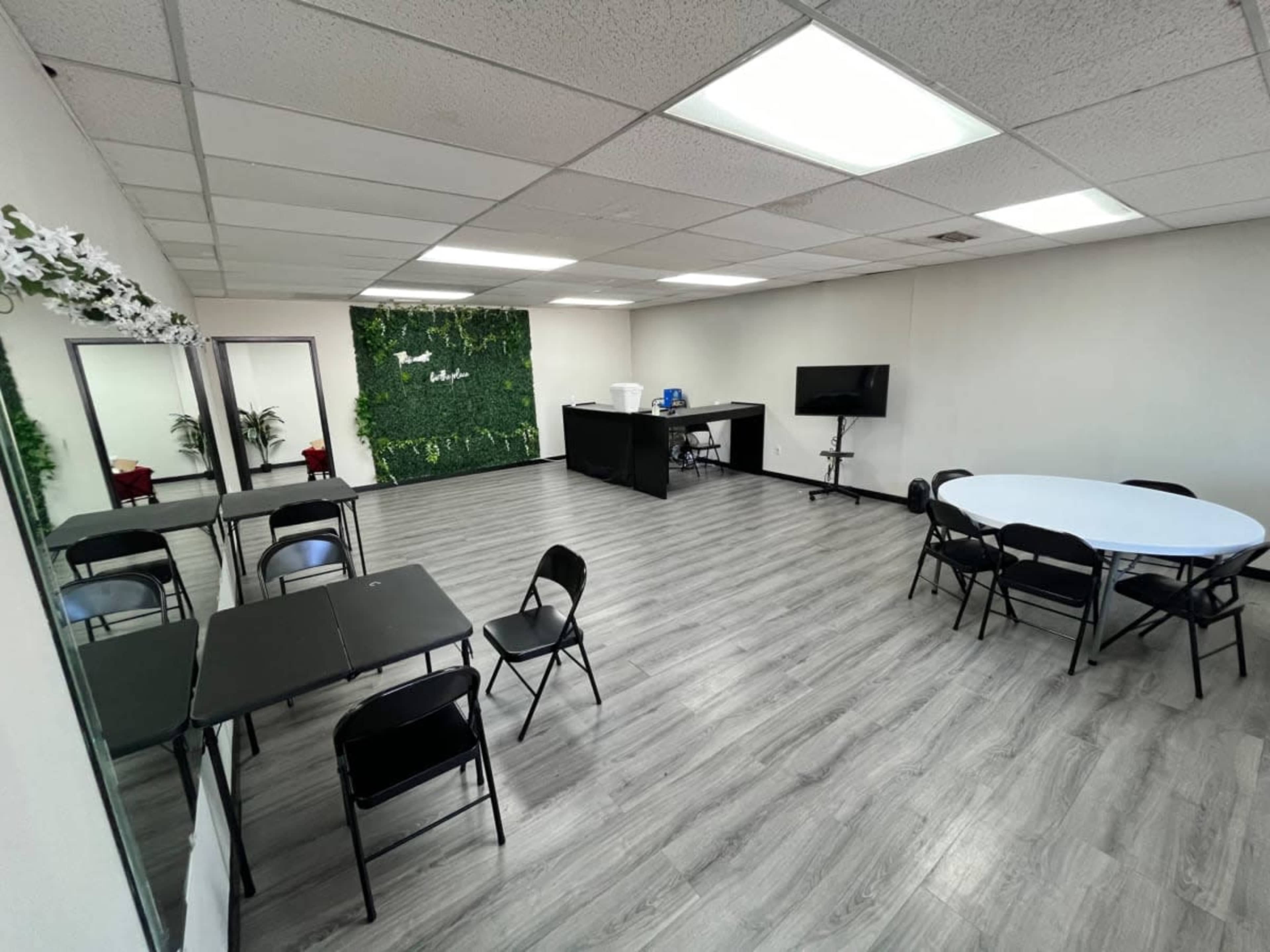 The image shows a spacious meeting room with tables and chairs arranged, a green plant wall, and a television setup.