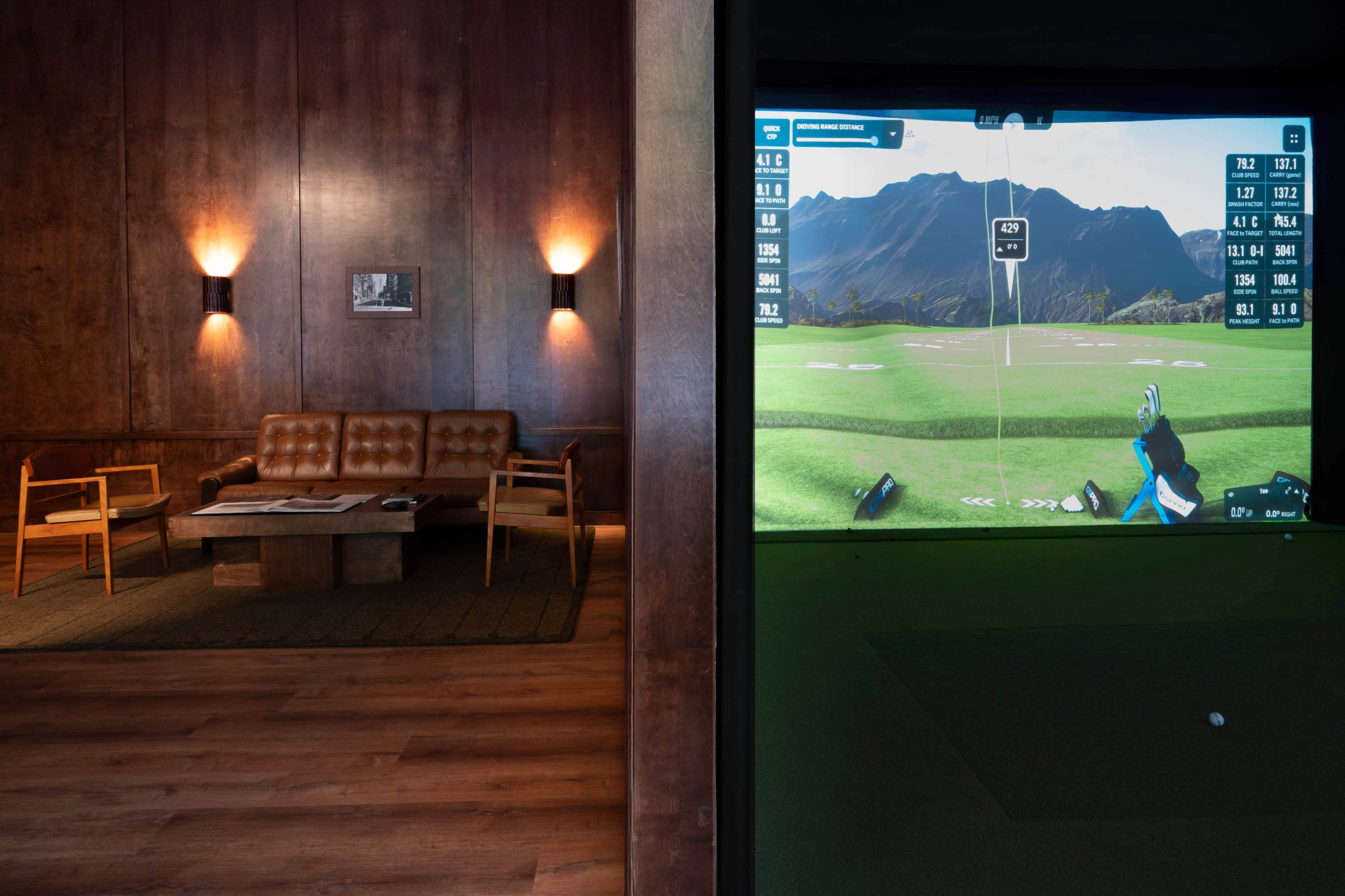 The image shows a stylish indoor space featuring a leather sofa and wooden furnishings on one side, while the other side displays a golf simulator with a mountainous landscape on the screen.