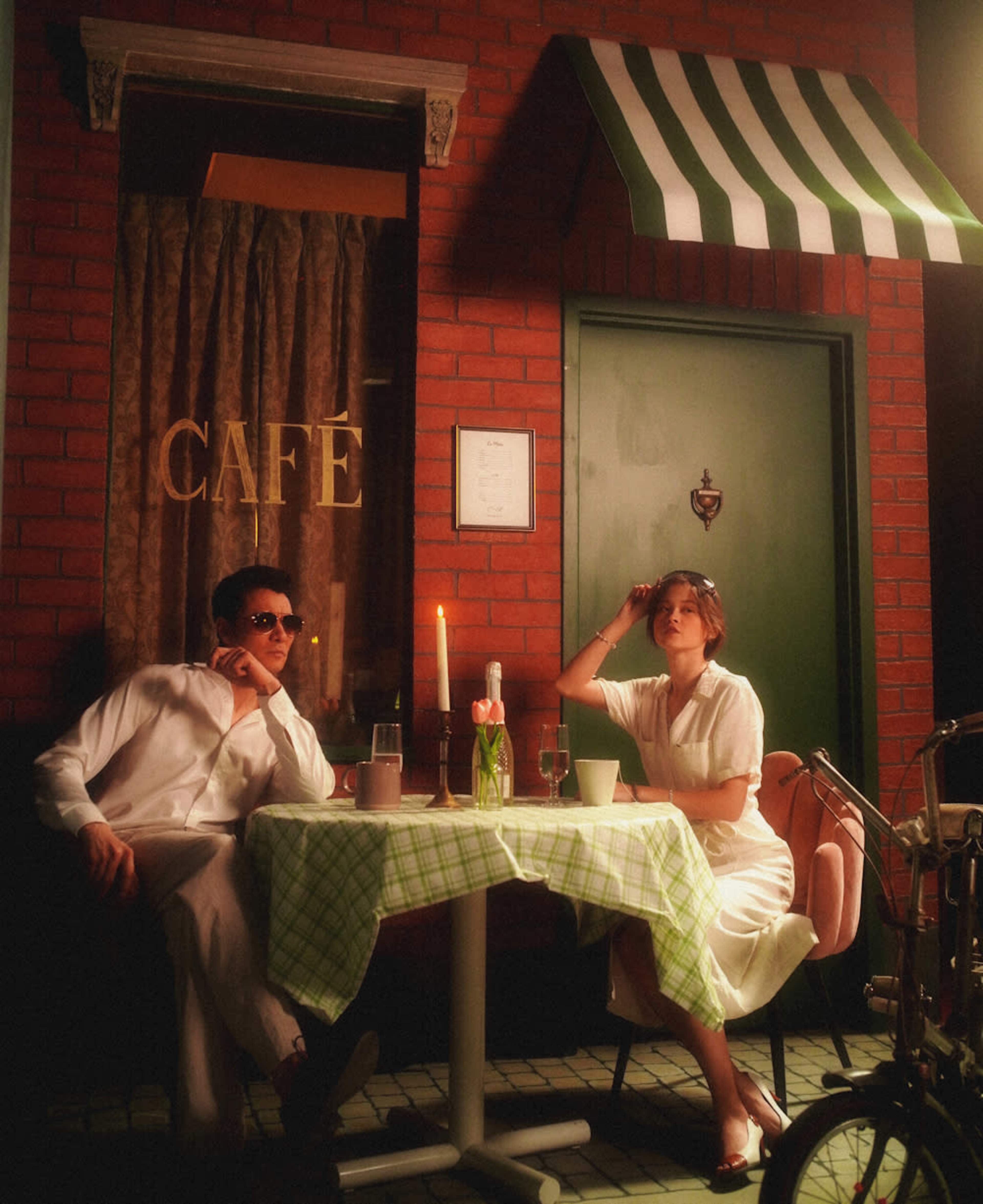 A man and a woman sit at a table outside a café, surrounded by brick walls and green accents, with a candle and drinks on the table.